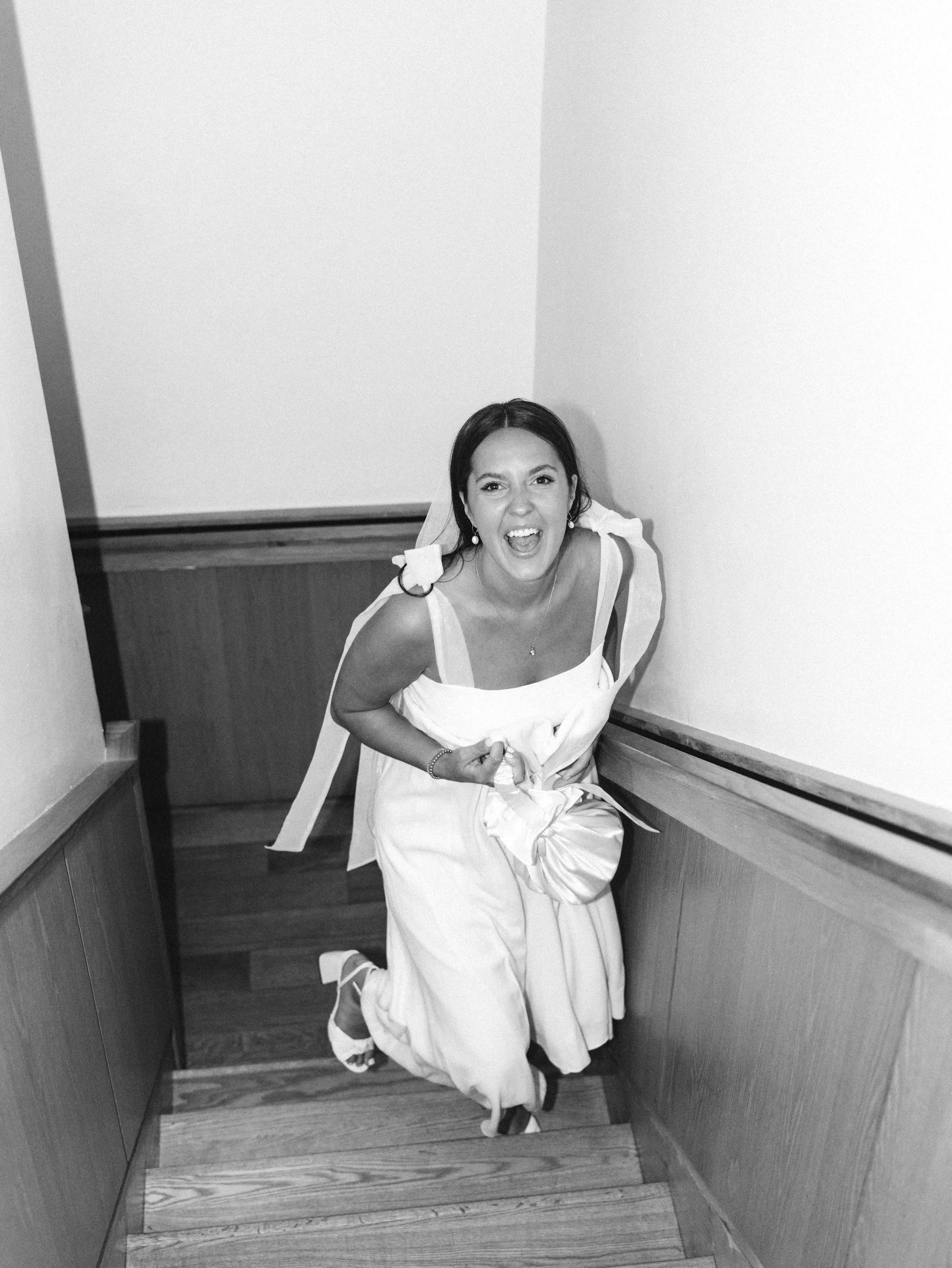 Woman in a white dress on a staircase, smiling widely and looking up at the camera.