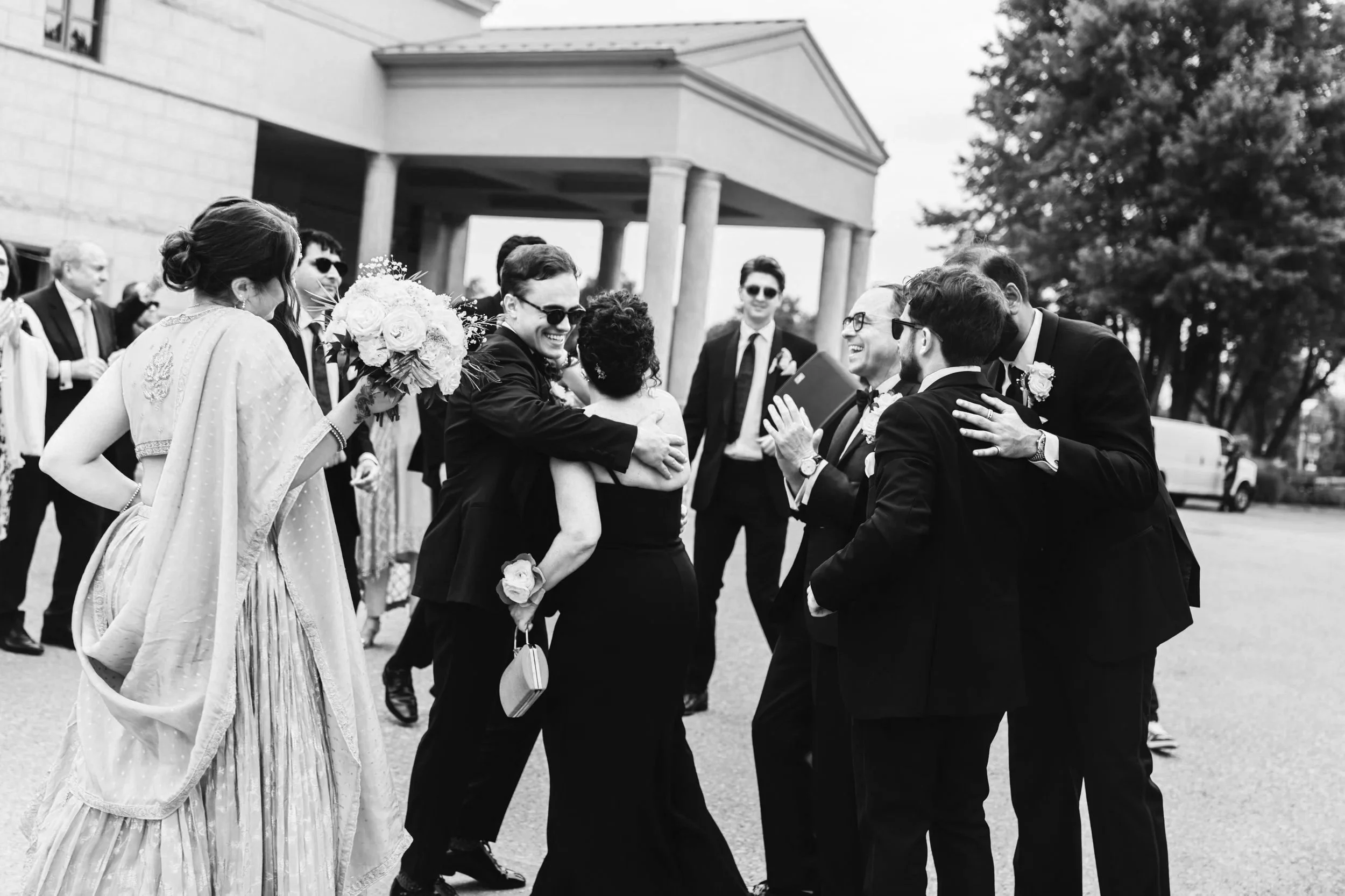 A group of people in formal attire celebrating outside a building, hugging and smiling, with wedding bouquets and glasses in hand.