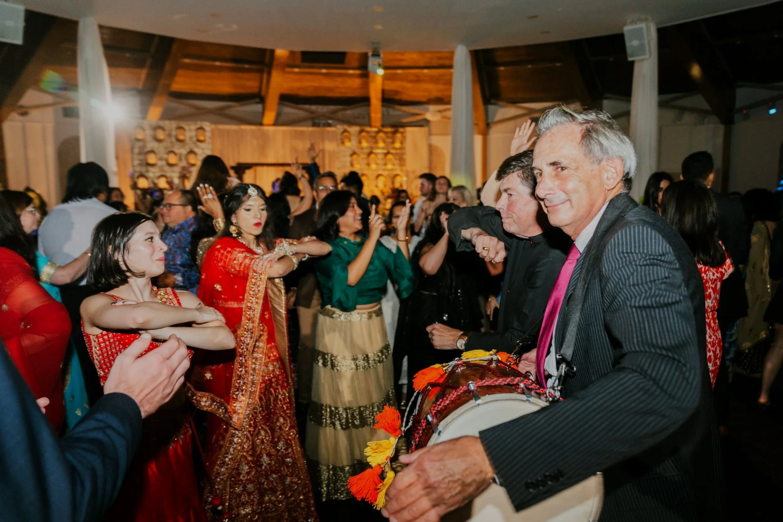 People dancing and celebrating at an event, with some dressed in traditional Indian attire. A man in the foreground is playing a drum with a smile on his face. The background shows a crowd enjoying the festivities.