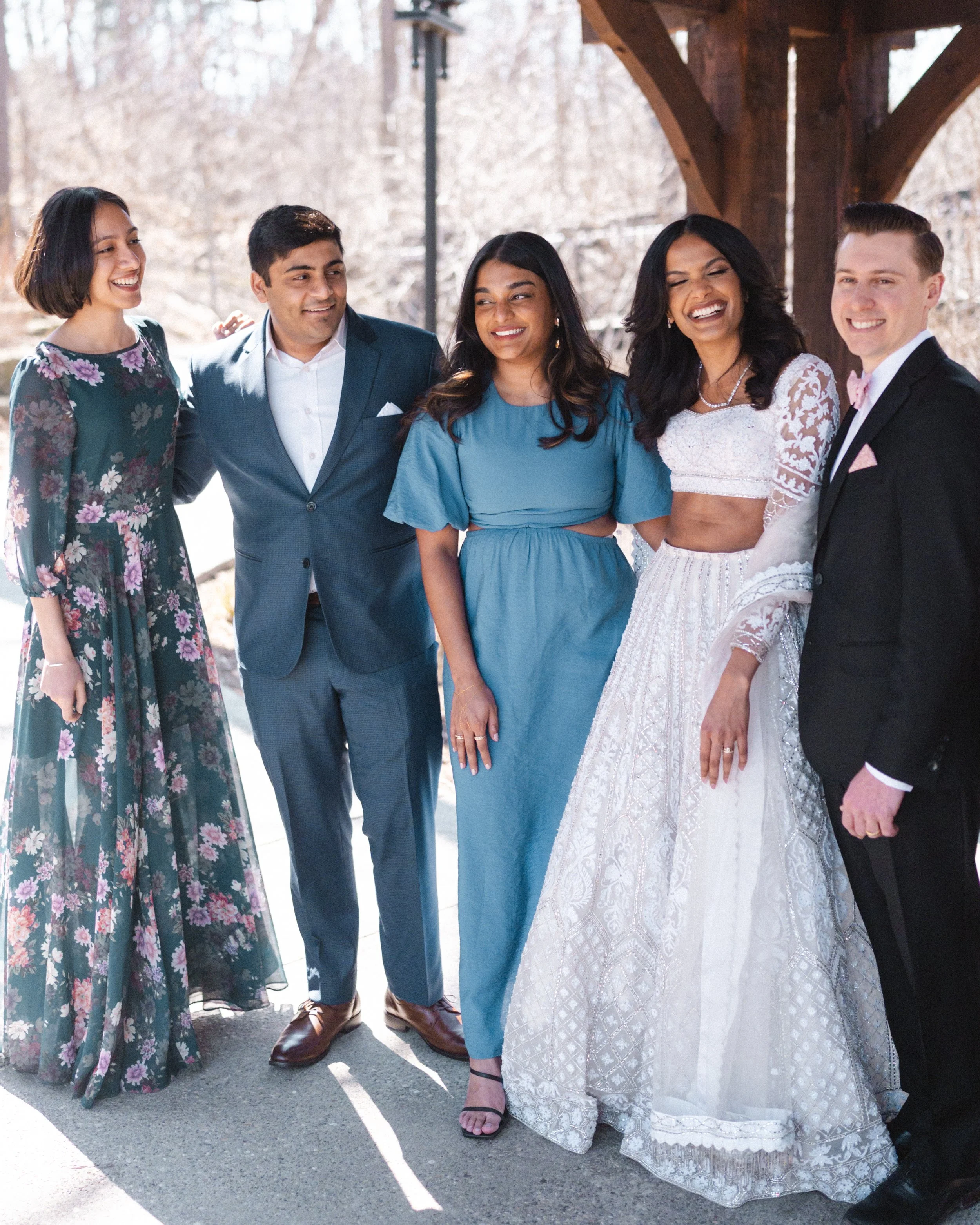 Group of six people standing outdoors in front of a wooden pavilion, dressed in formal attire and smiling.