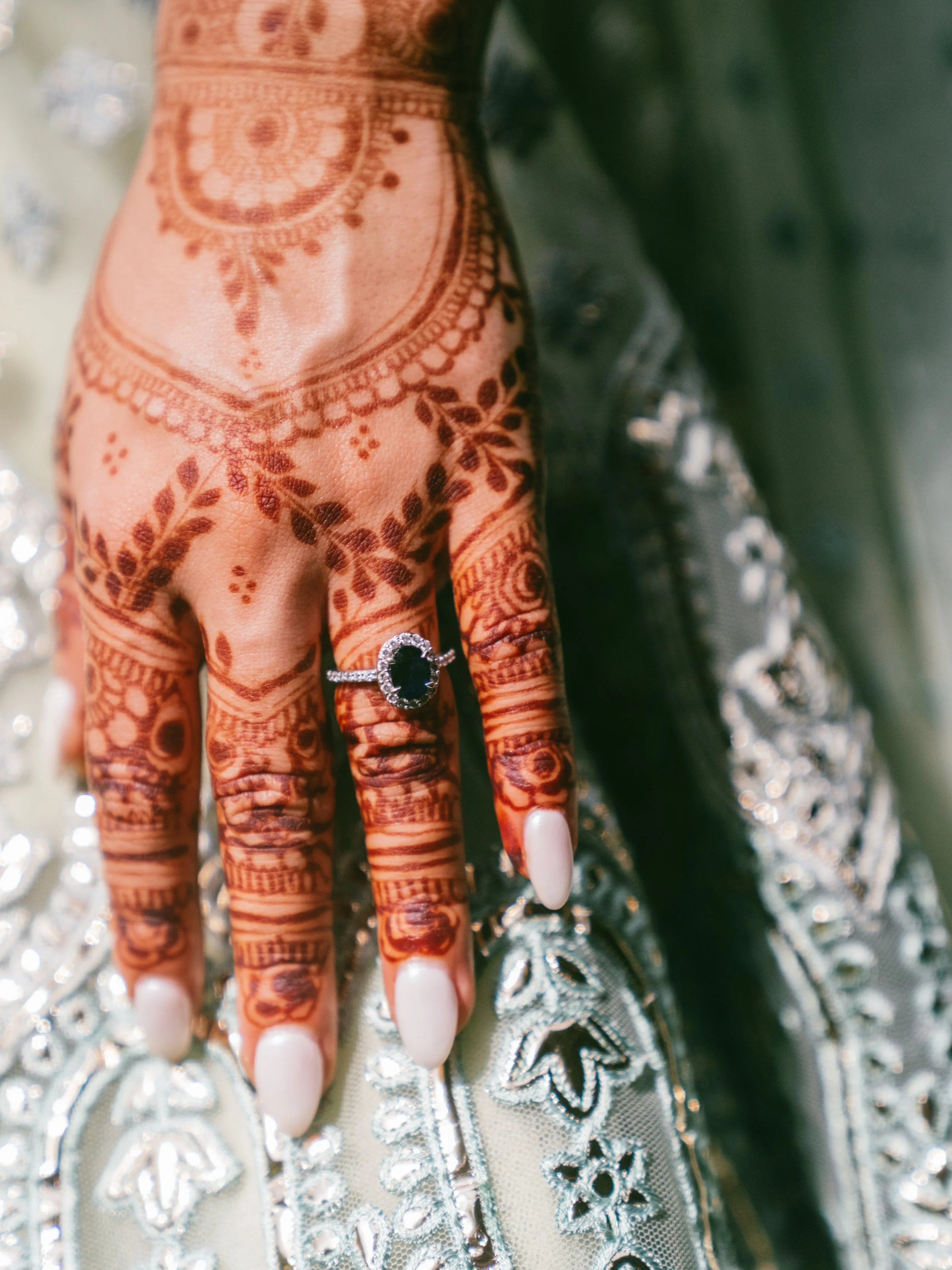 Close-up of a hand with henna tattoos, a silver ring with a black gemstone, and long white nails. The hand rests on a fabric with intricate silver embroidery.
