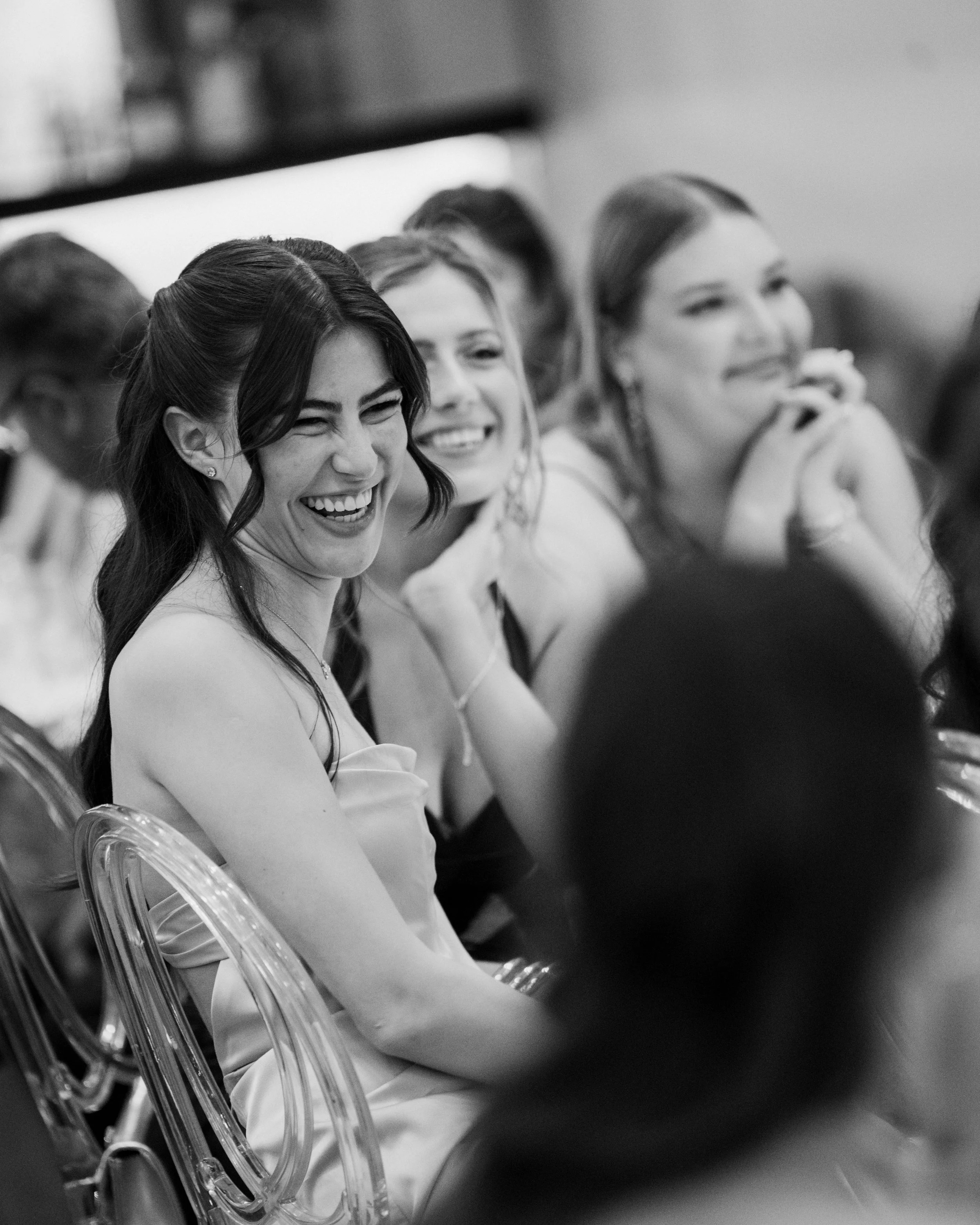 Group of women laughing and smiling at an event, seated in transparent chairs, in black and white photo.