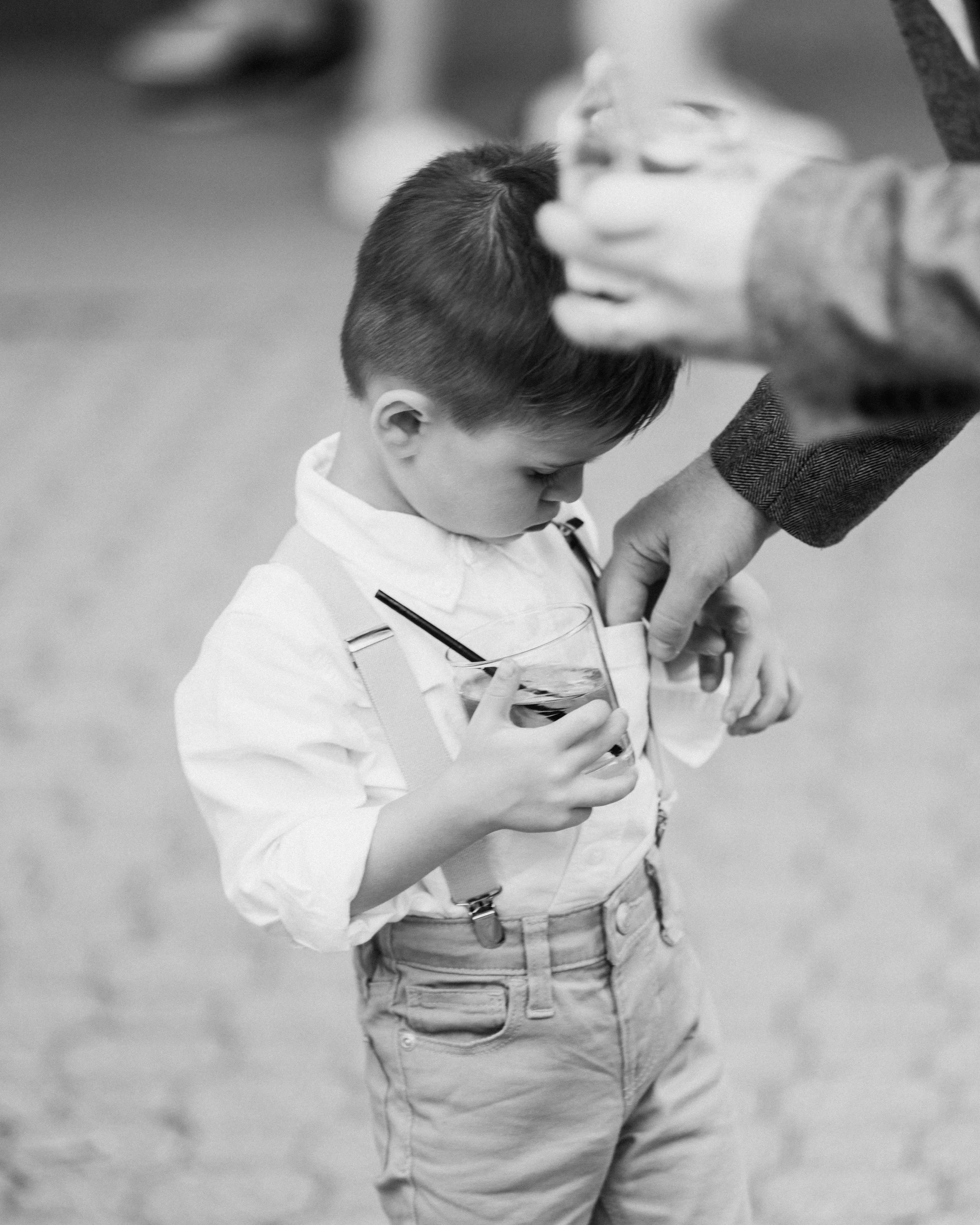 A young boy in a white shirt with suspenders holding a glass of soda with a straw, standing with his head bowed and looking into the glass, while an adult adjusts his shirt.