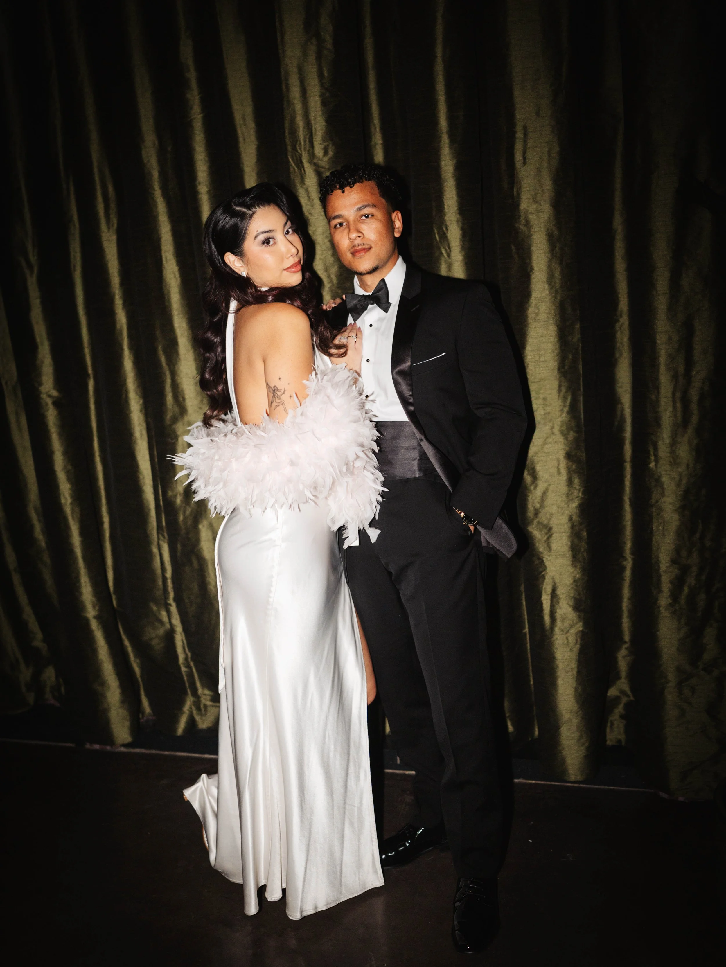 A man and woman dressed in formal attire posing in front of a dark curtain. The woman wears a white gown with feathered accessories, and the man wears a black tuxedo with a bow tie.