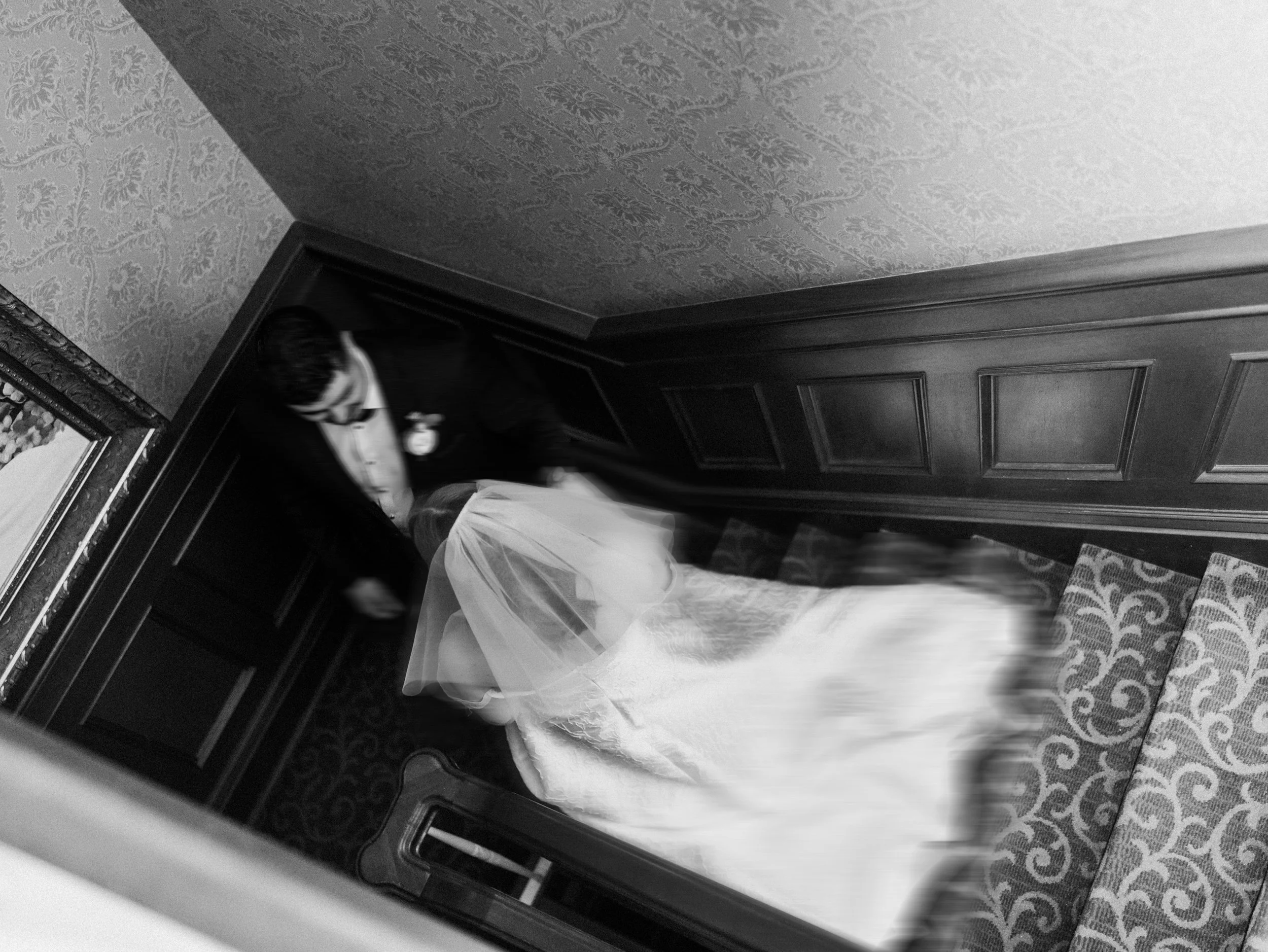 A man in a tuxedo, possibly a groom, helping a woman in a wedding dress, possibly a bride, down a staircase in an elegant, vintage-style interior.