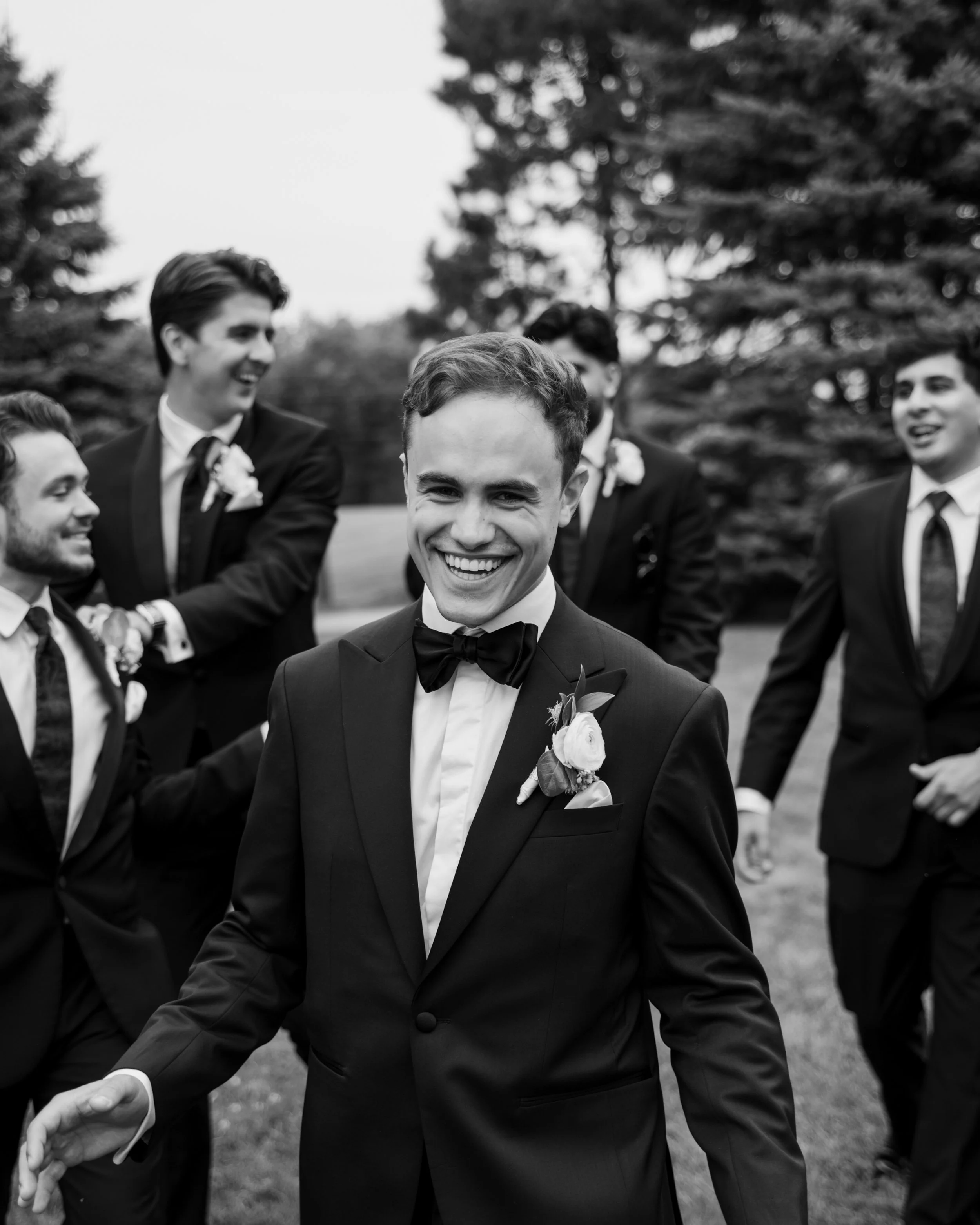 Black and white photo of a groom and his groomsmen outdoors, all in tuxedos, smiling and walking.