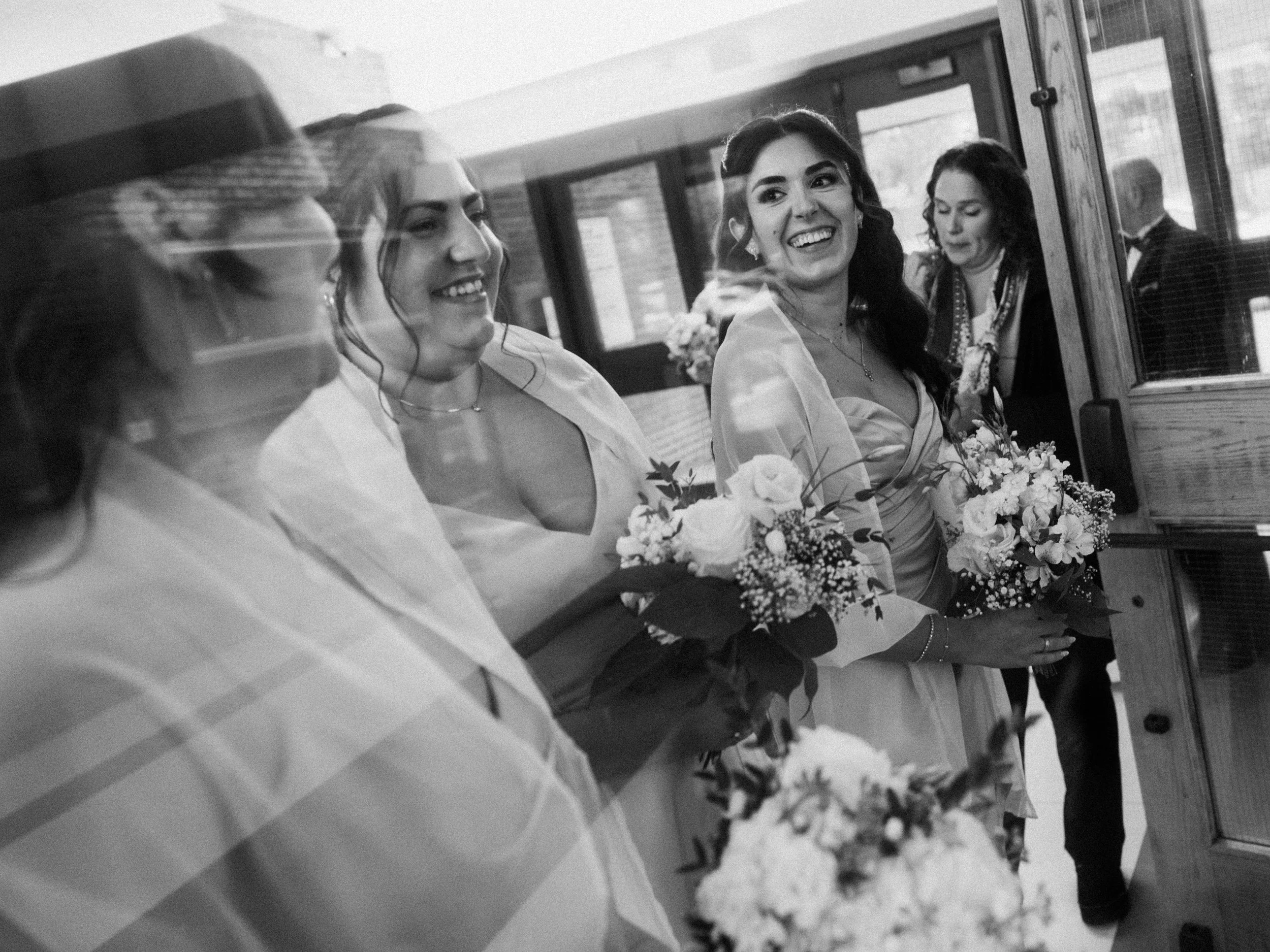 Group of women dressed in formal attire holding bouquets, smiling and standing inside a vintage train or trolley.