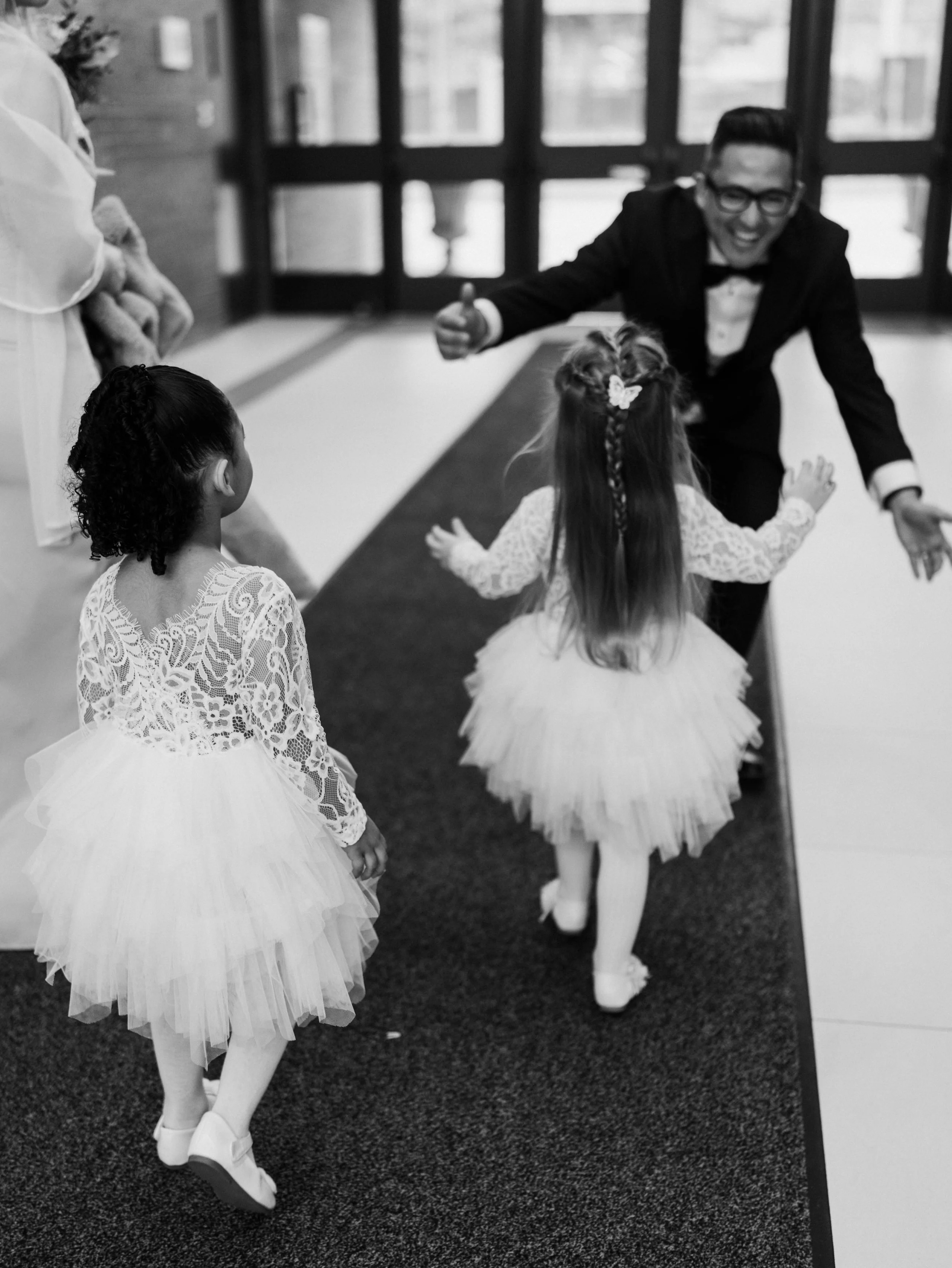 A man in a tuxedo and glasses is joyfully running towards two young girls in fancy dresses inside a building with glass doors in the background.