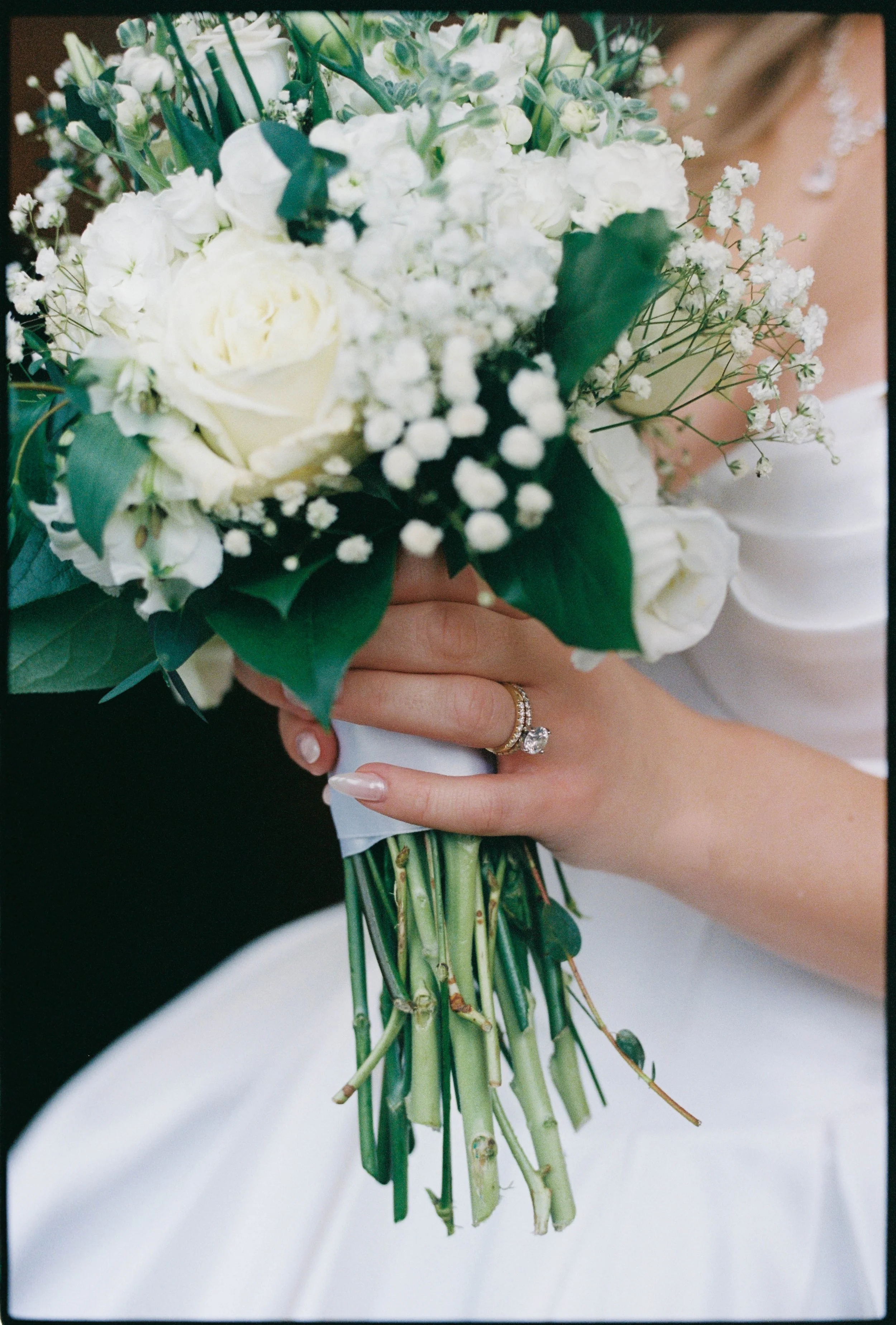 Close-up of a bride's hand holding a bouquet of white flowers, including roses and baby's breath, with a diamond engagement ring and wedding band, against a blurred background.