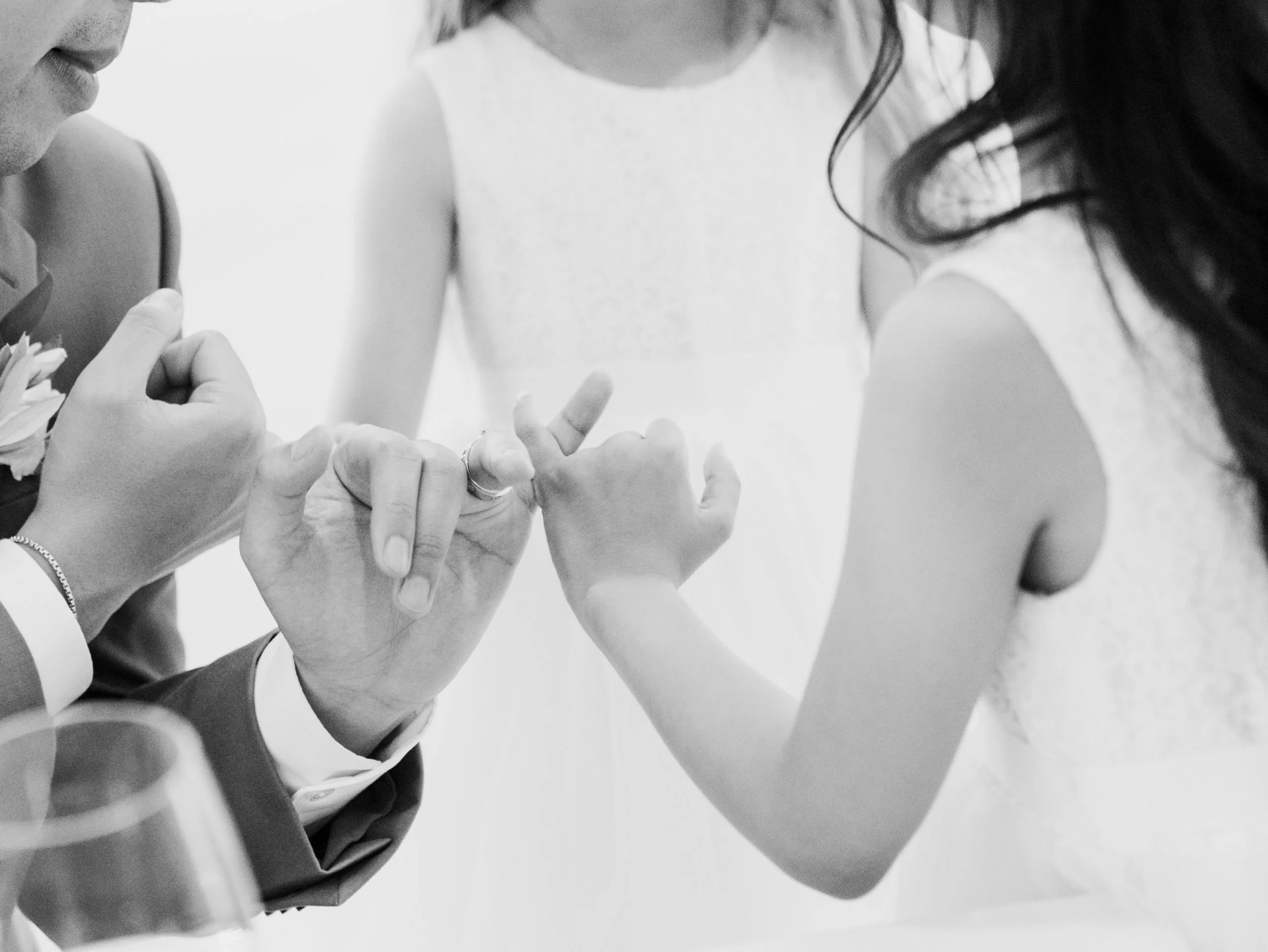 A couple exchanging rings during a wedding ceremony, with a person in a white dress standing nearby.