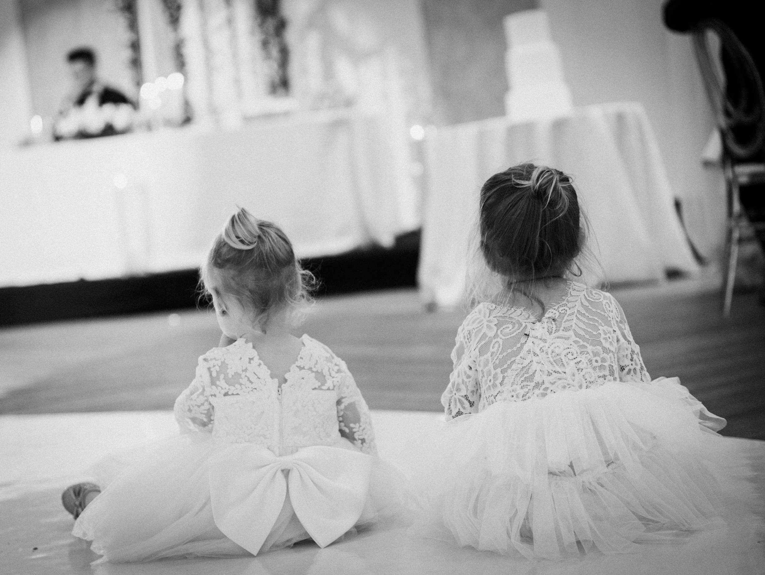 Two young girls in lace dresses sitting on the floor at an indoor event, facing away from the camera, with a blurred background including a woman at a table and a large decorated cake.