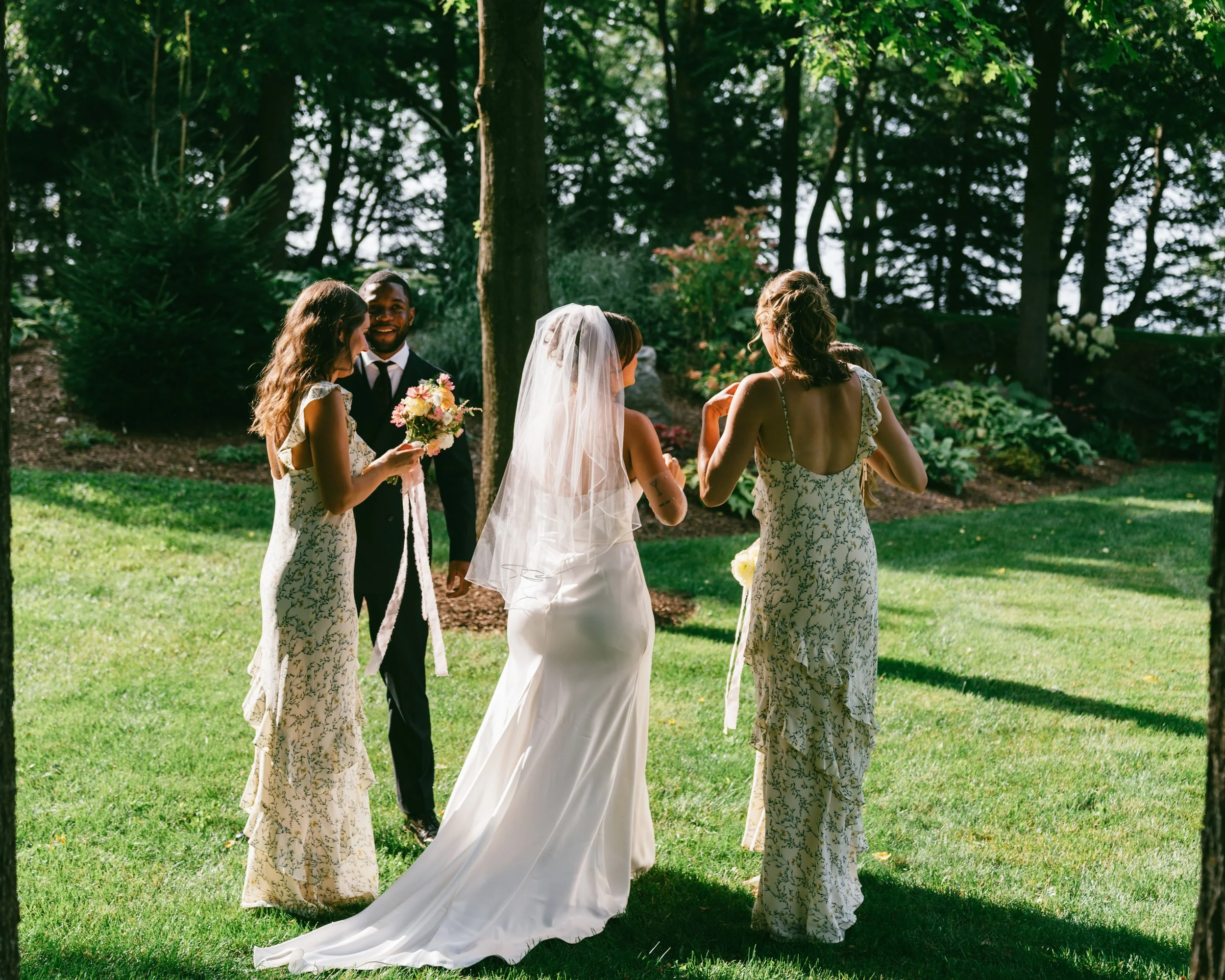 A wedding ceremony outdoors with a bride in a white gown and veil standing with four people, three women and one man, and a garden with trees and bushes in the background.