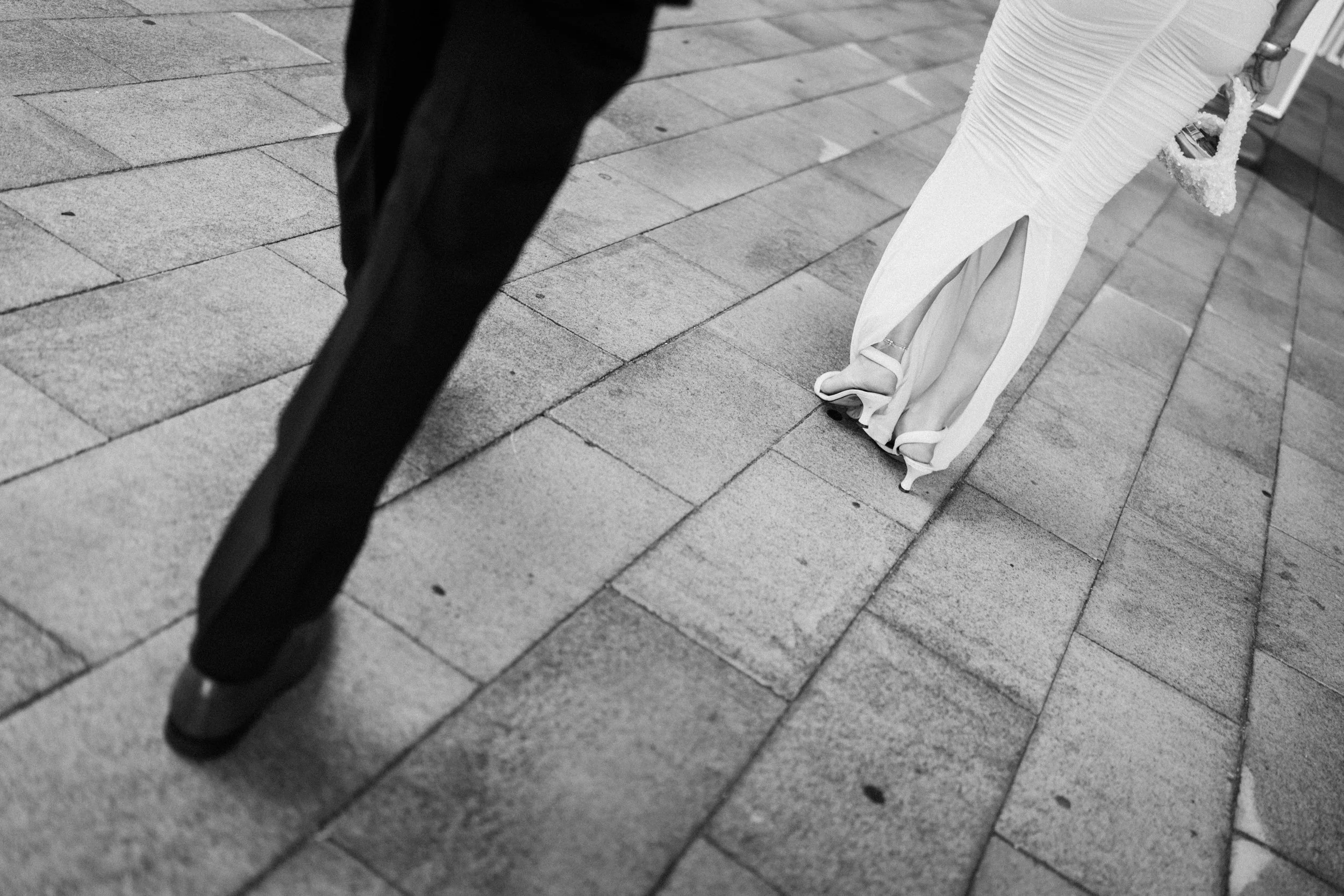 A black and white photo showing a person in dark pants and shoes walking on a brick sidewalk next to a woman in a long white dress and high heels.