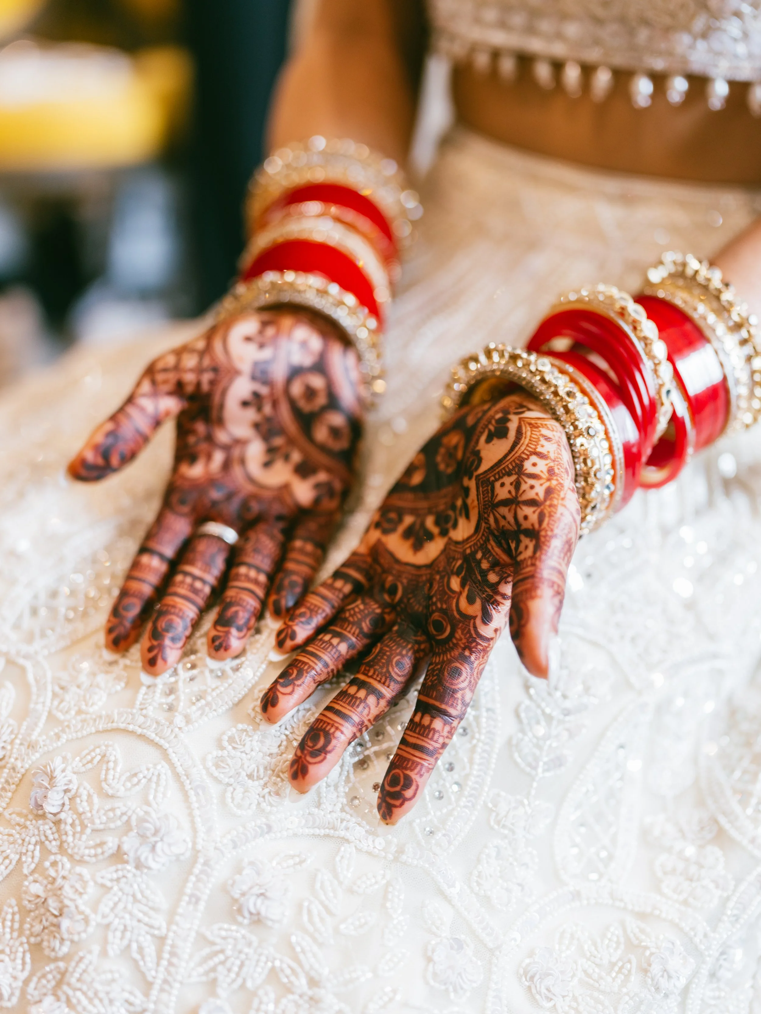 Close-up of a woman's decorated hands with traditional henna designs, red and gold bangles, and ornate clothing, likely for a wedding or cultural celebration.