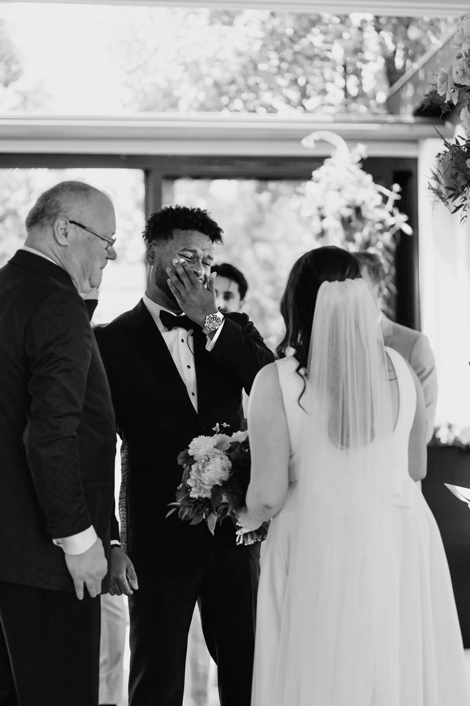 Black and white photo of a wedding ceremony with a joyful groom wiping tears, bride holding a bouquet, and officiant and another person present.