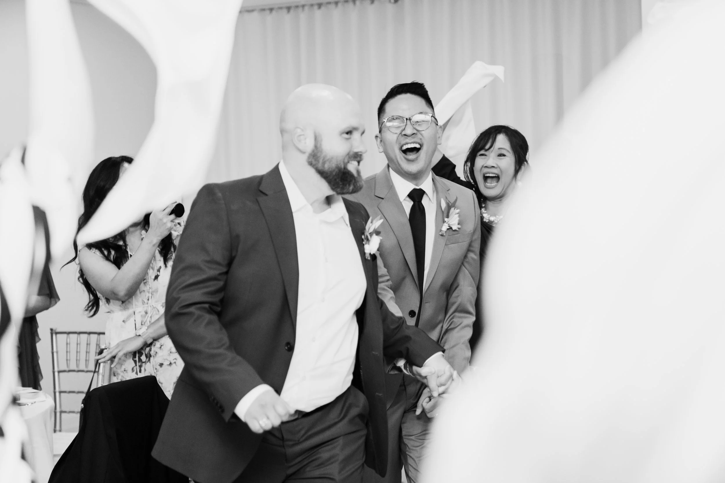 Black and white photo of wedding celebration with people smiling and dancing, holding hands, surrounded by paper decorations.