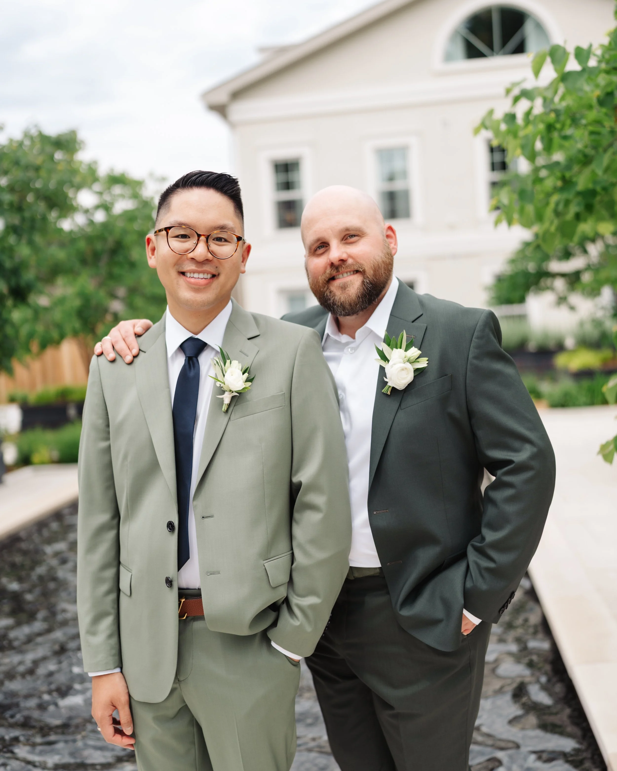 Two men in suits with boutonnières standing outdoors in front of a white house with trees, smiling, one has glasses, the other is bald with a beard.