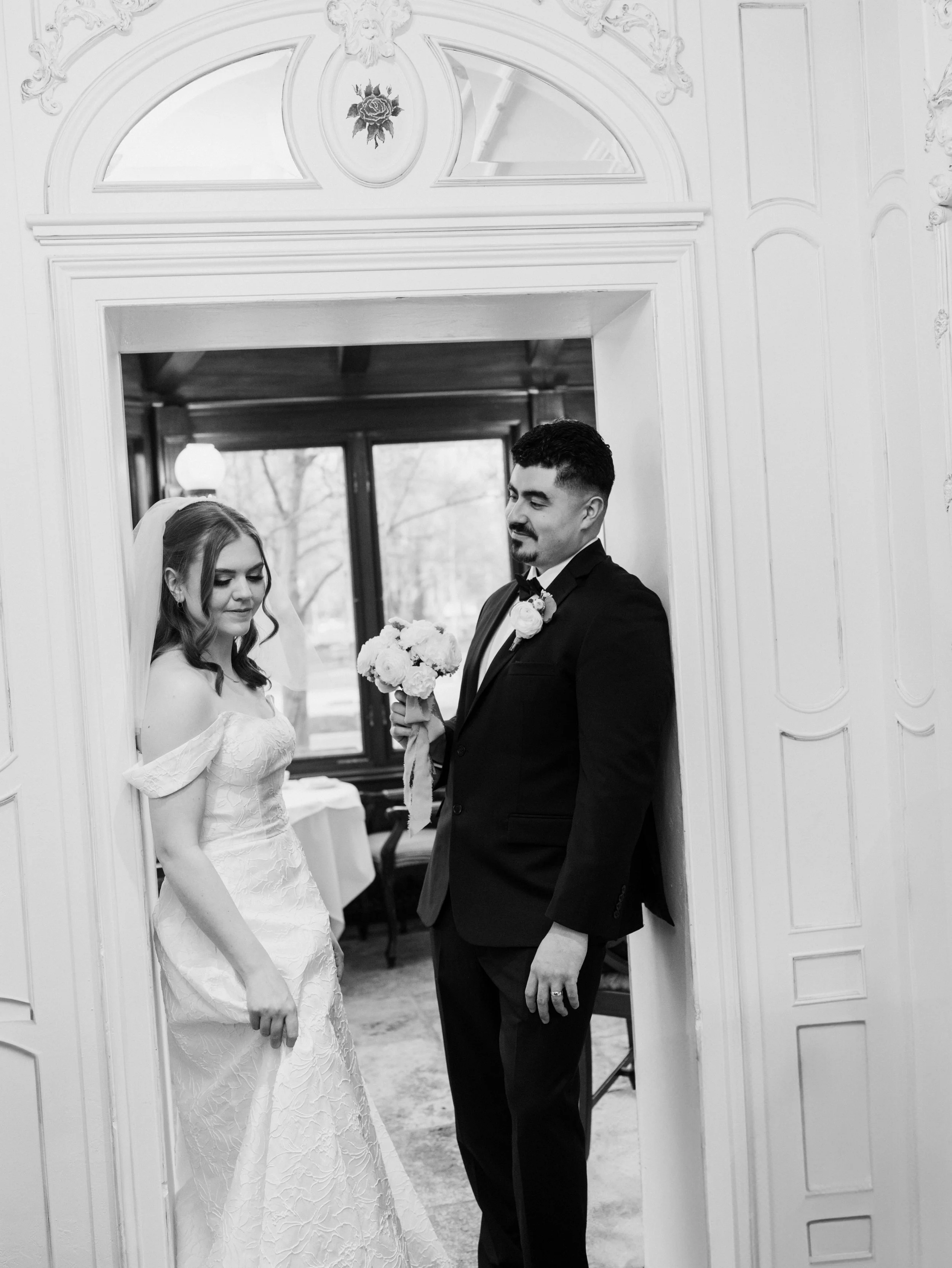 A black and white photo of a bride and groom inside a decorated room, with the groom offering a bouquet to the bride.