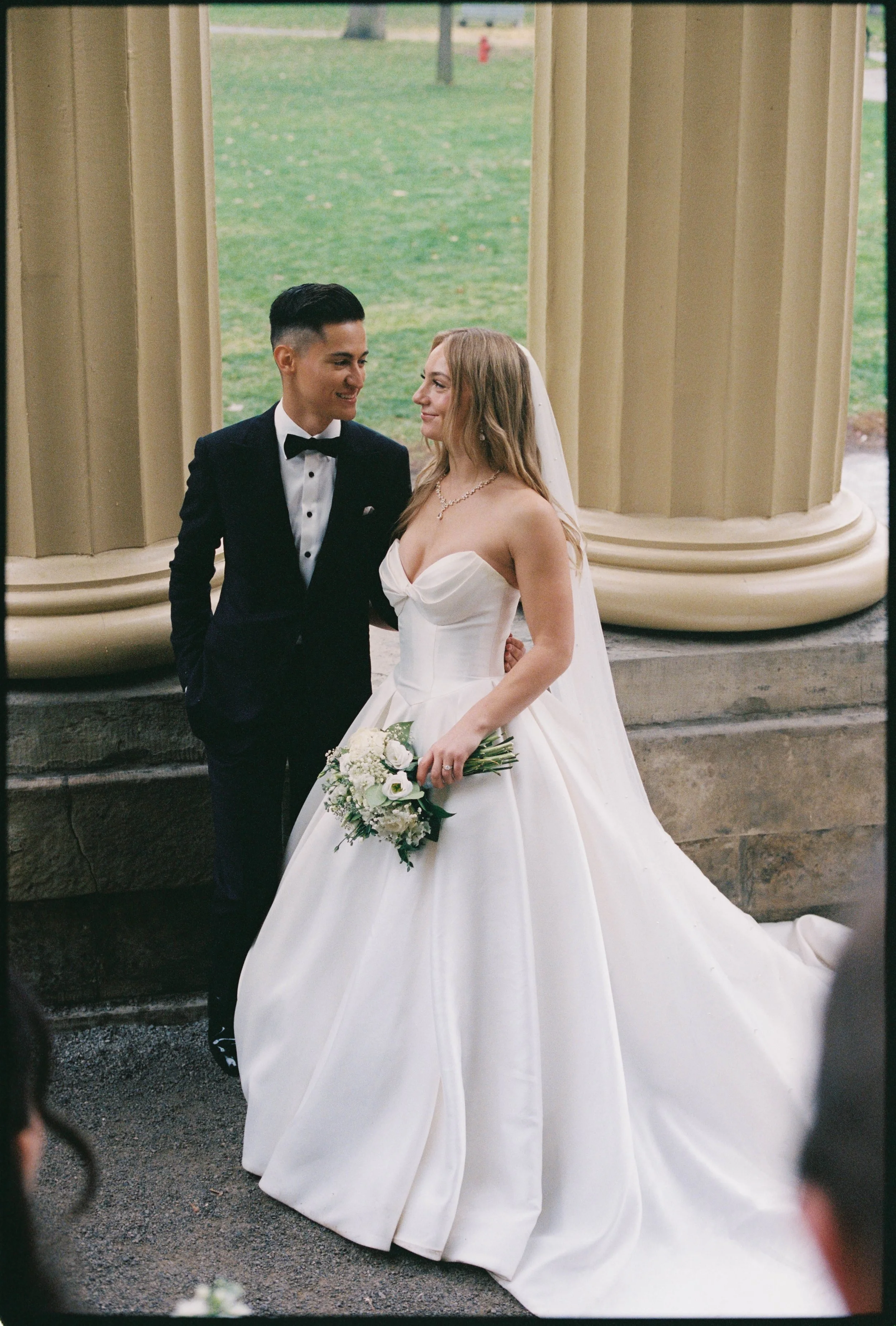 A bride and groom standing close together during their wedding ceremony, with the bride holding a bouquet of white flowers. The groom is wearing a black tuxedo with a bow tie, and the bride is wearing a strapless white wedding gown with a veil.