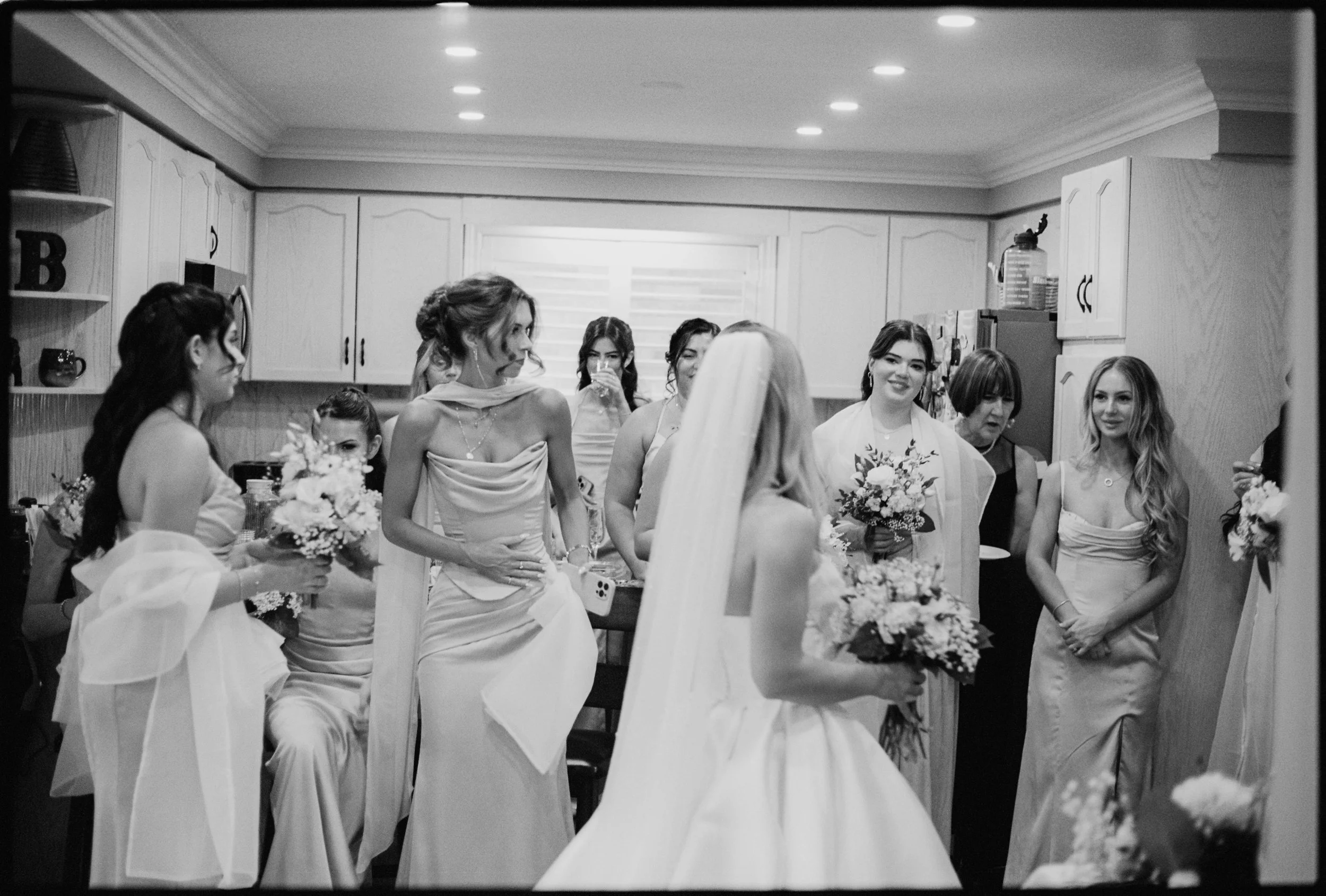 A black and white photo of a bride in a wedding dress holding a bouquet. She is surrounded by women, likely bridesmaids, in a kitchen with white cabinets, some holding bouquets, all smiling and looking at the bride.