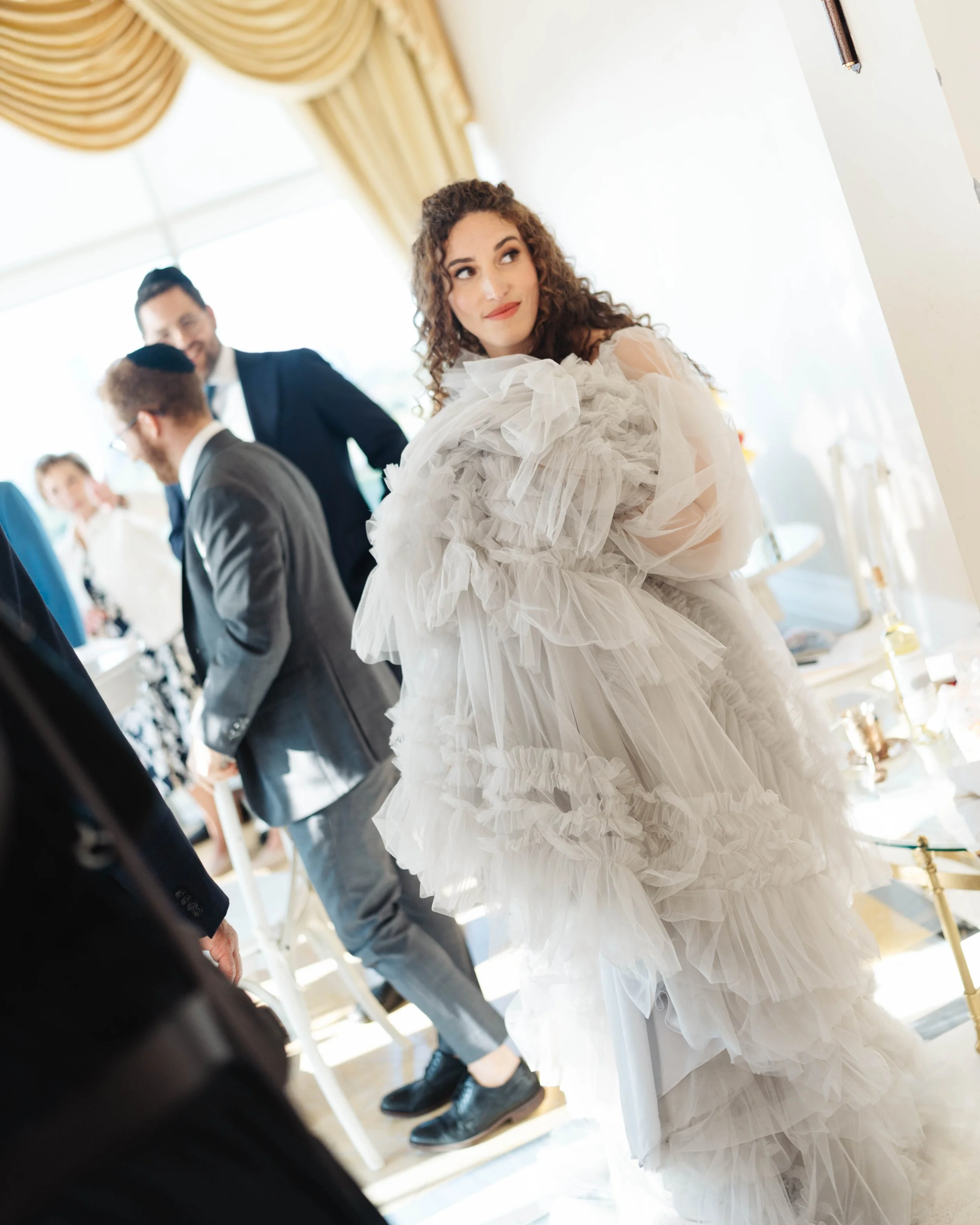 Woman in a ruffled white dress at a social gathering, with men in suits in the background.