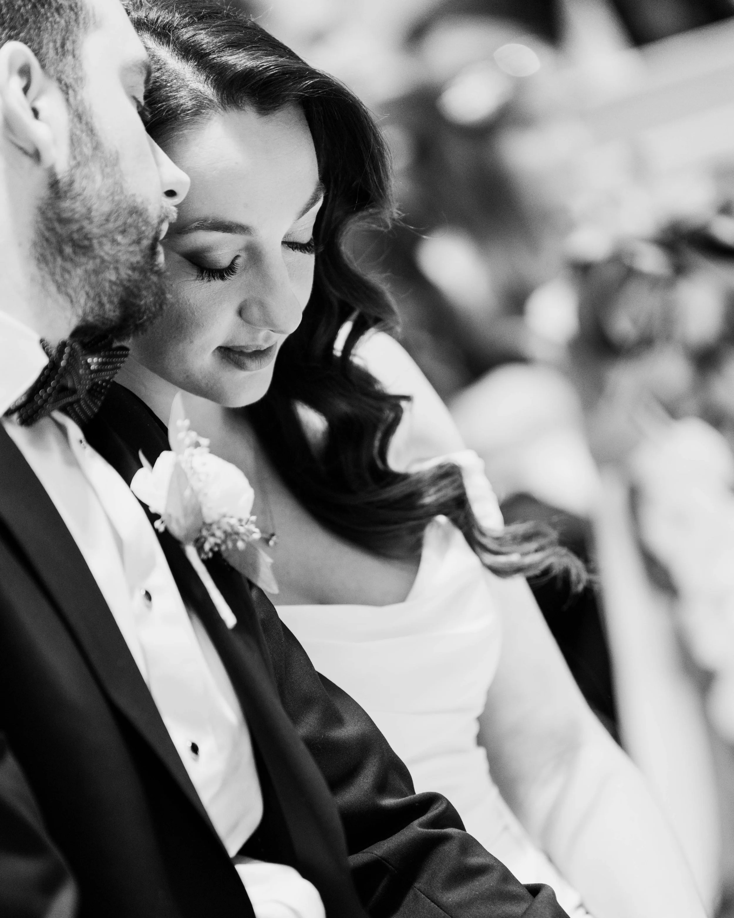 Black and white photo of a bride and groom in a close-up, with their eyes closed, touching foreheads, dressed in wedding attire.