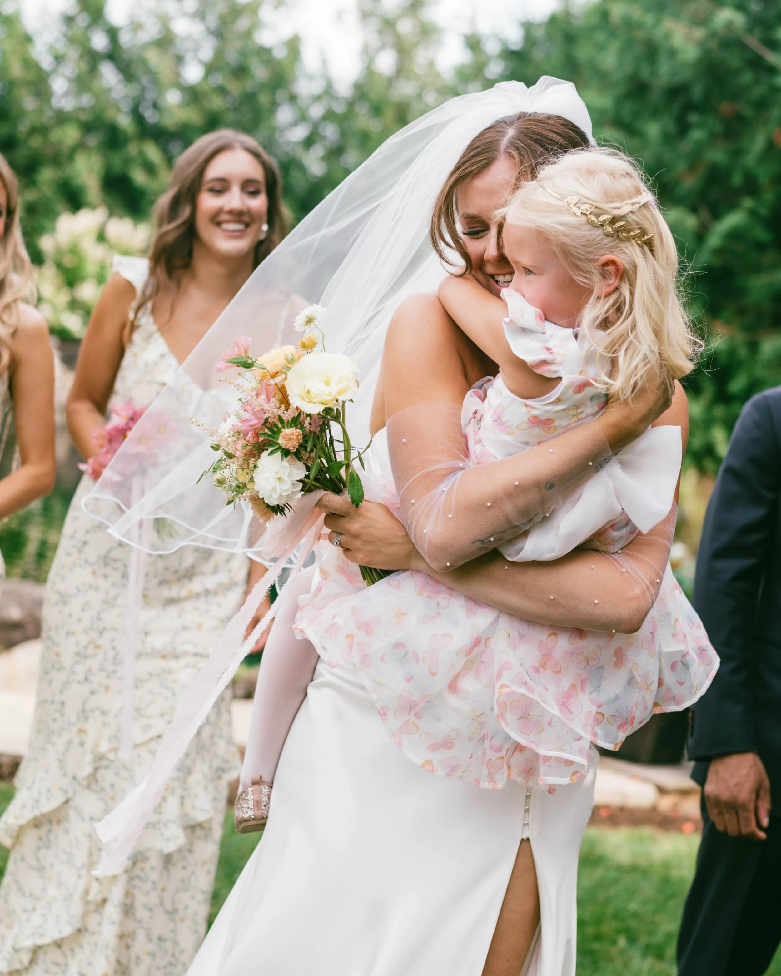 A bride in a white wedding dress hugging a young girl in a floral dress, with a wedding bouquet, while smiling and surrounded by friends outdoors.