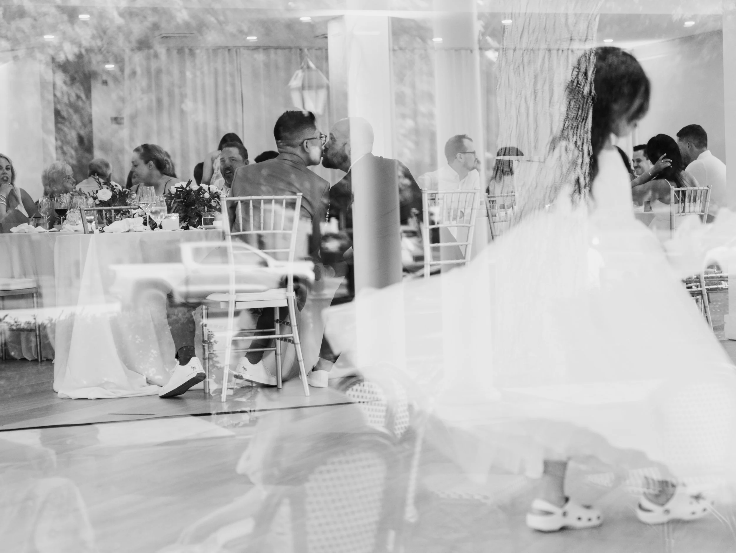 Black and white photograph of a wedding reception seen through a glass window. Guests are sitting at decorated tables, talking and enjoying the celebration. The reflection of the street and car outside is visible on the glass.