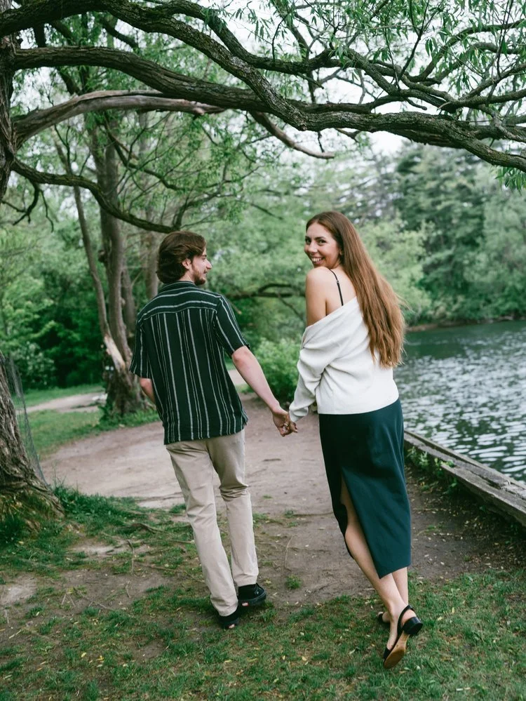 A man and woman holding hands walking by a lake in a wooded area, smiling and looking back at the camera.