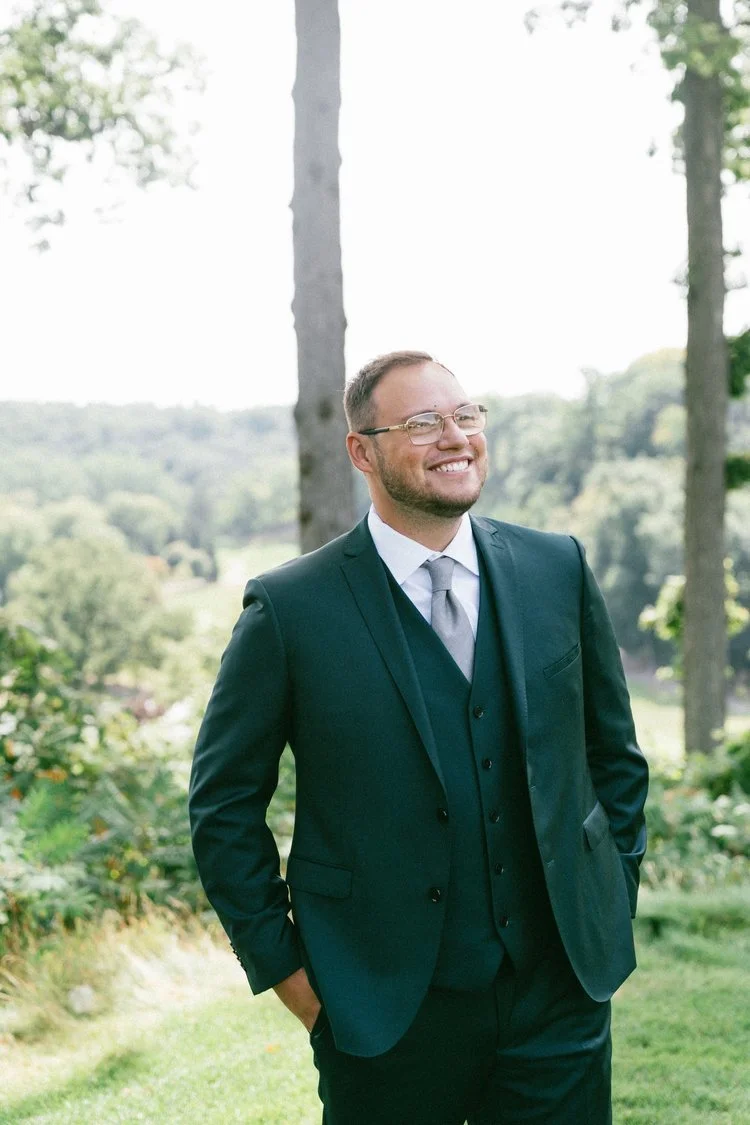 A groom at his outdoor Wedding ceremony in a green suit, smiling outdoors at his bride walking down the aisle