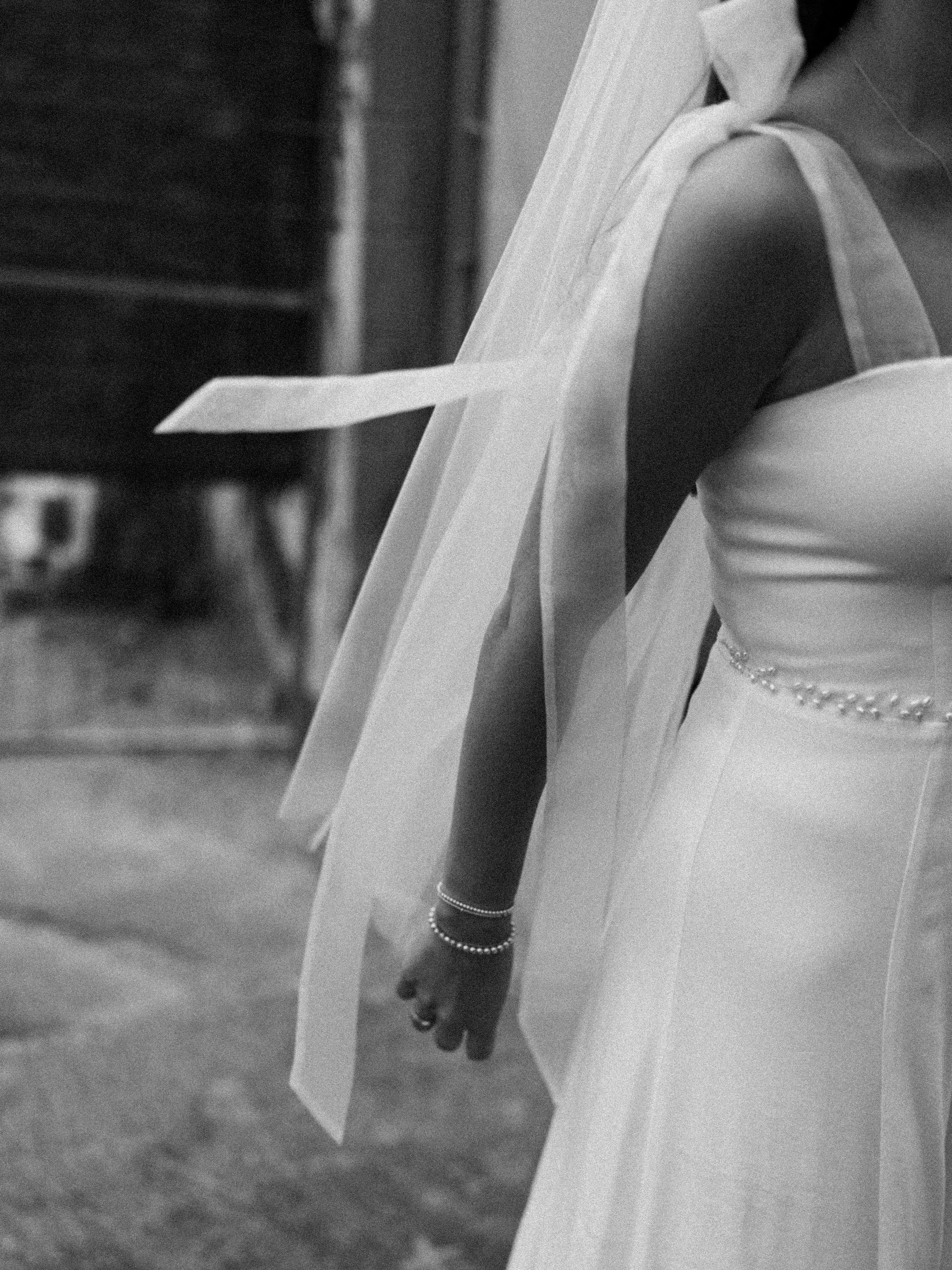 Close-up of a bride in a wedding dress, showing her arm, wrist jewelry, and part of her veil in black and white.