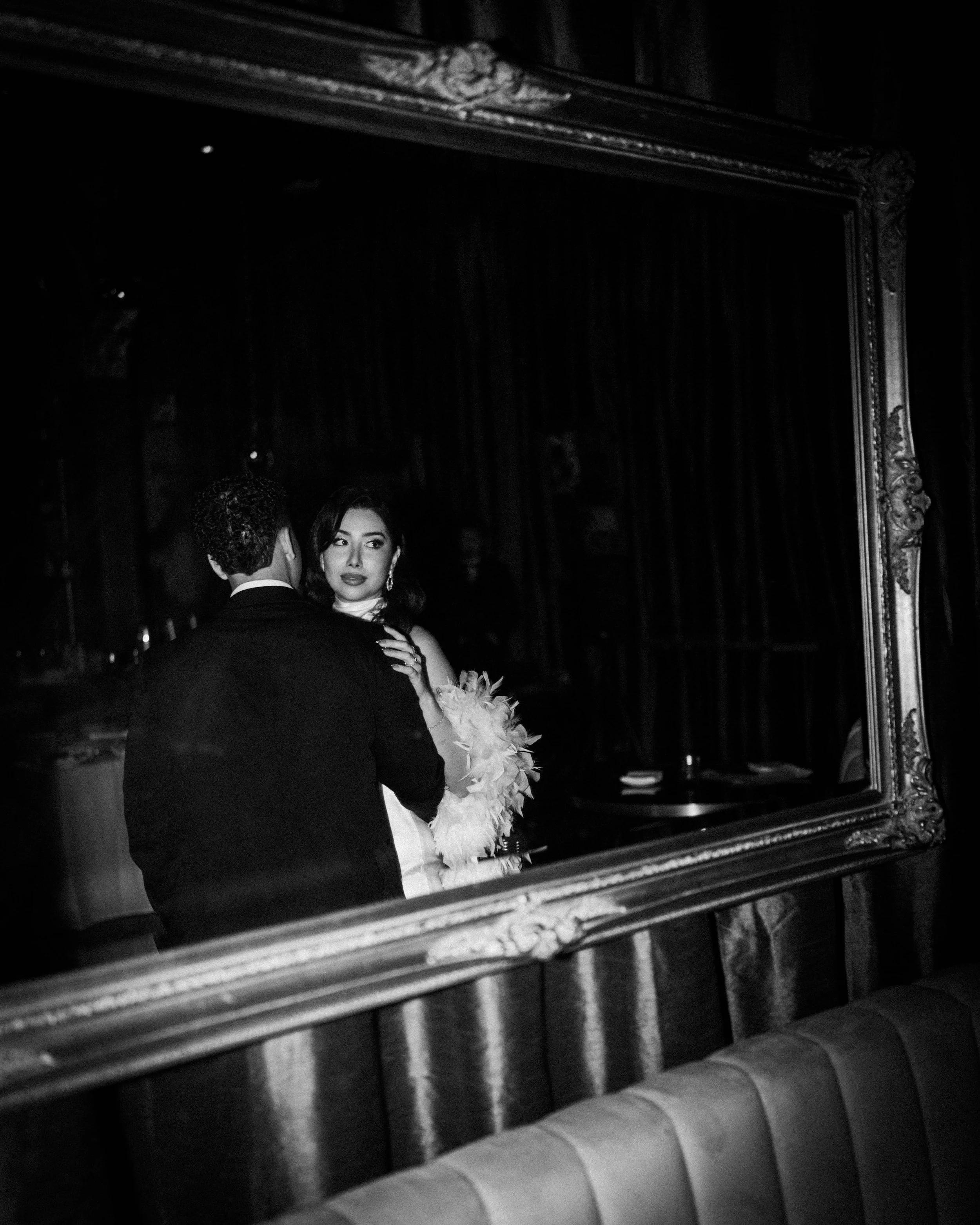 A black and white photo of a man and woman dancing in front of a large mirror, with the woman's face visible and the man’s back turned. The woman is holding a feathered fan and wearing elegant earrings.