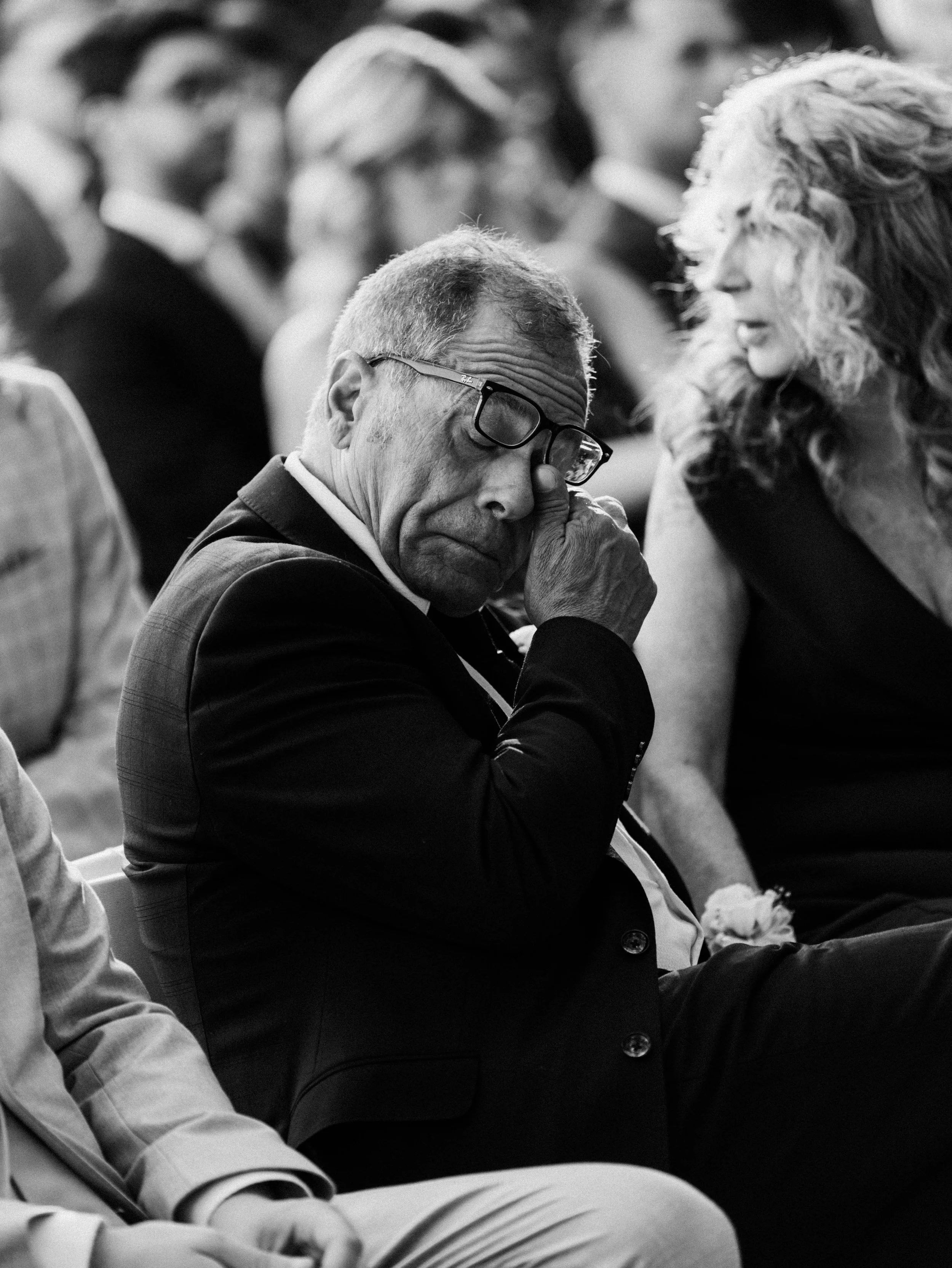 A man wearing glasses and a suit is sitting in a crowded space, with his hand raised to his face, appearing emotional or distressed. A woman next to him is speaking to him, with a visible expression of concern or empathy.