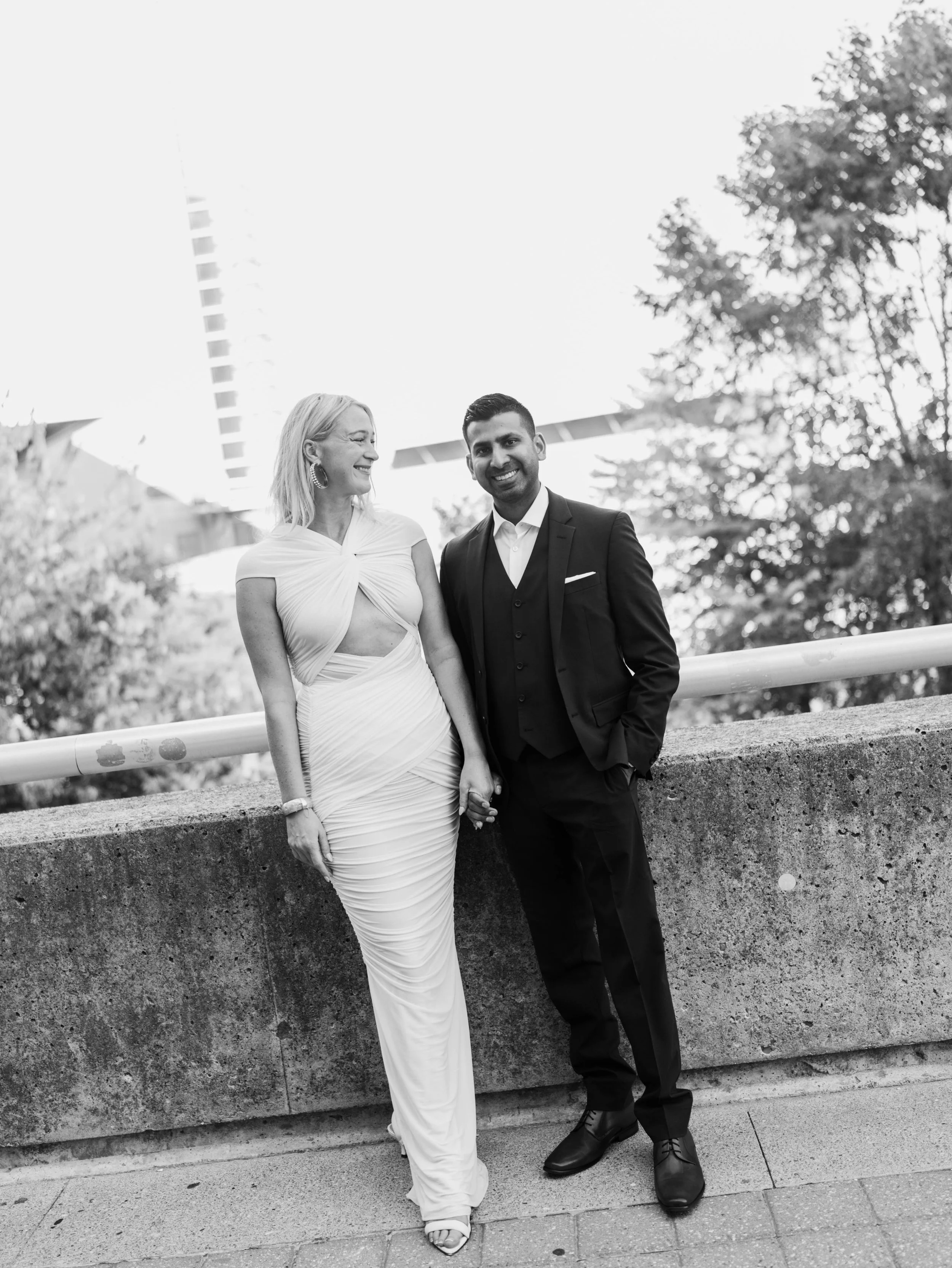 A smiling woman in a fashionable, elegant dress holding hands with a man in a tuxedo, posing outdoors on a city street with trees and modern buildings in the background.