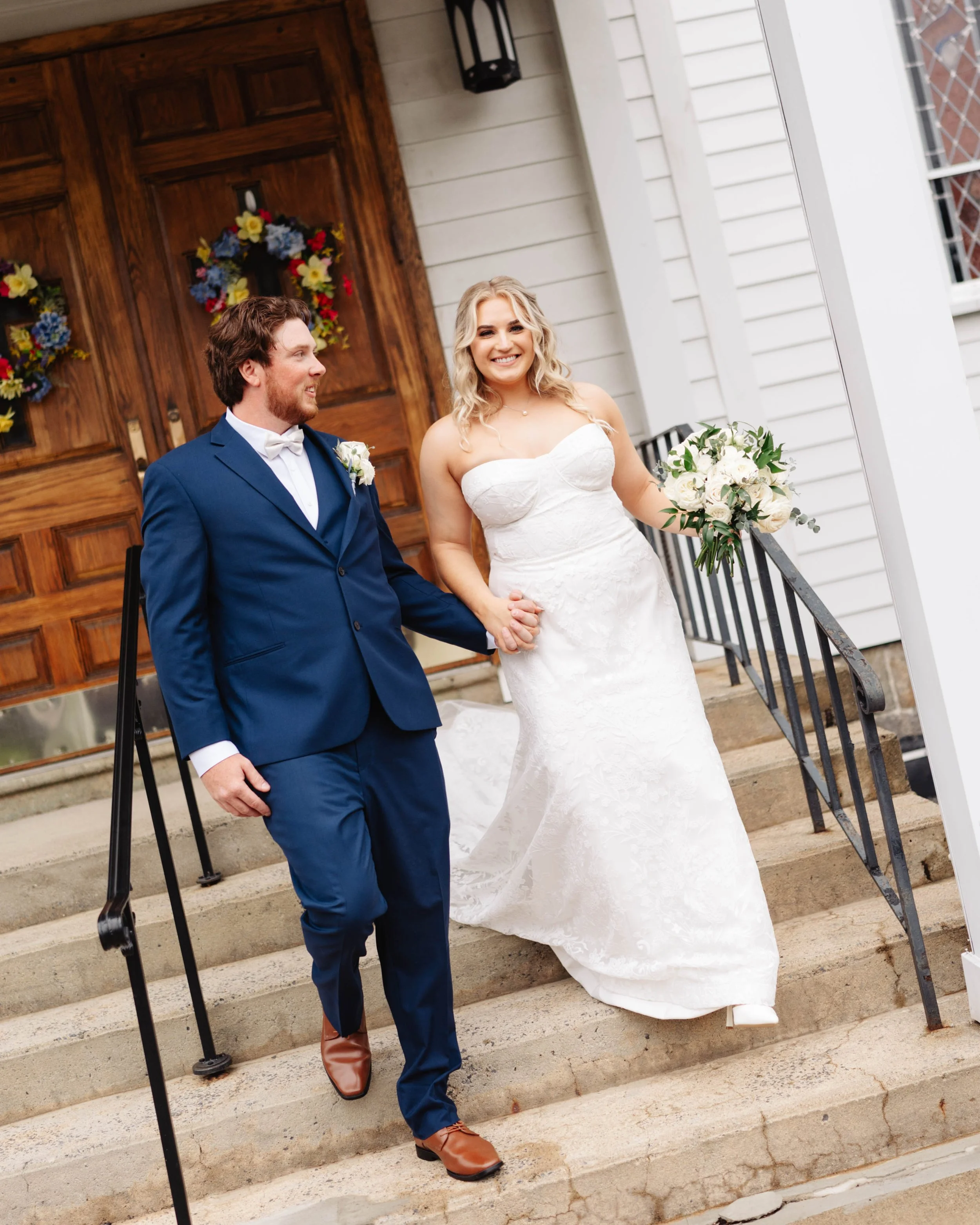 A bride and groom holding hands and smiling as they walk down the steps outside a house decorated for a wedding.