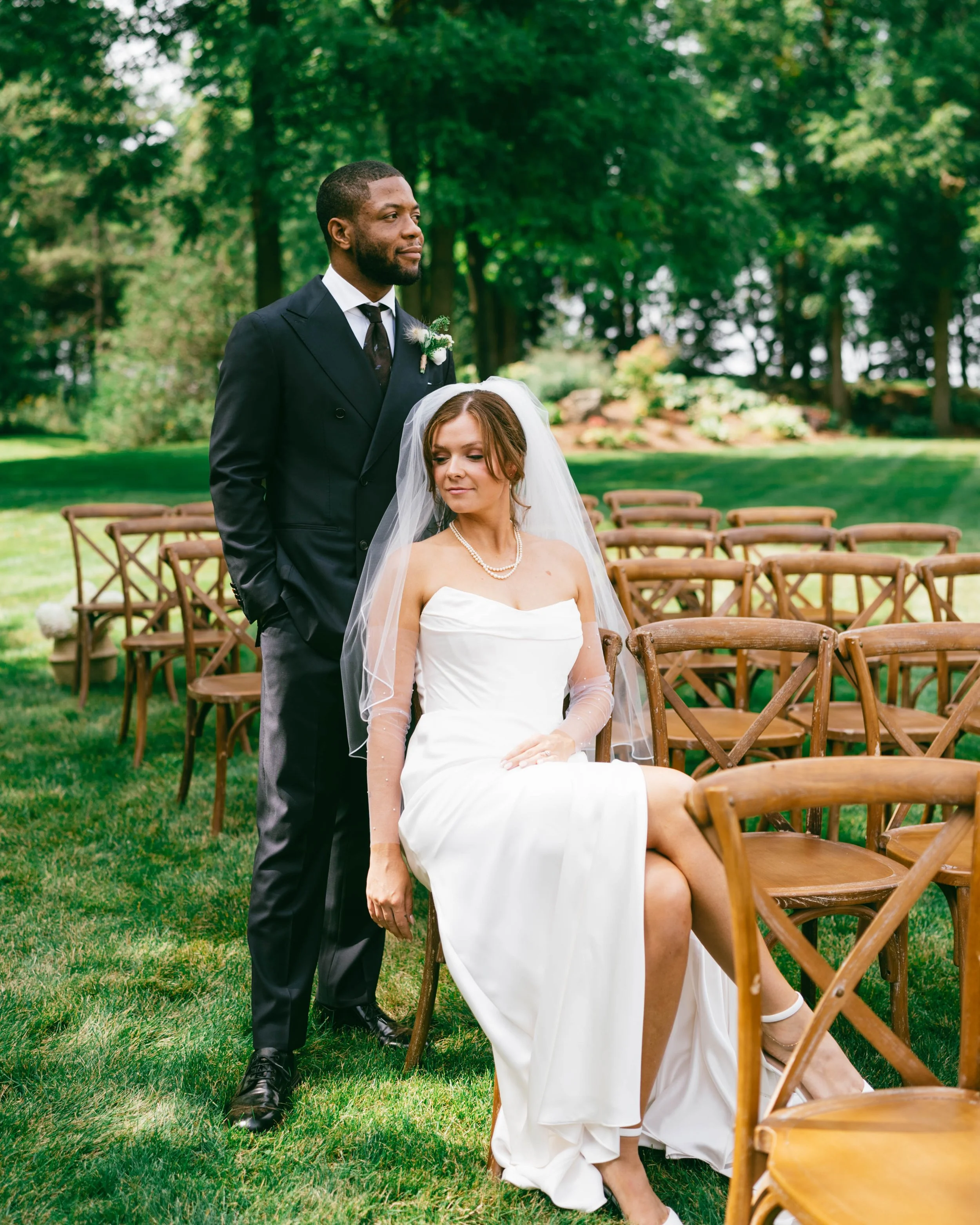 A bride in a white strapless wedding gown and veil sitting on a wooden chair outdoors, with a groom in a black suit and tie standing behind her. They are in a lush green garden with multiple empty chairs around.