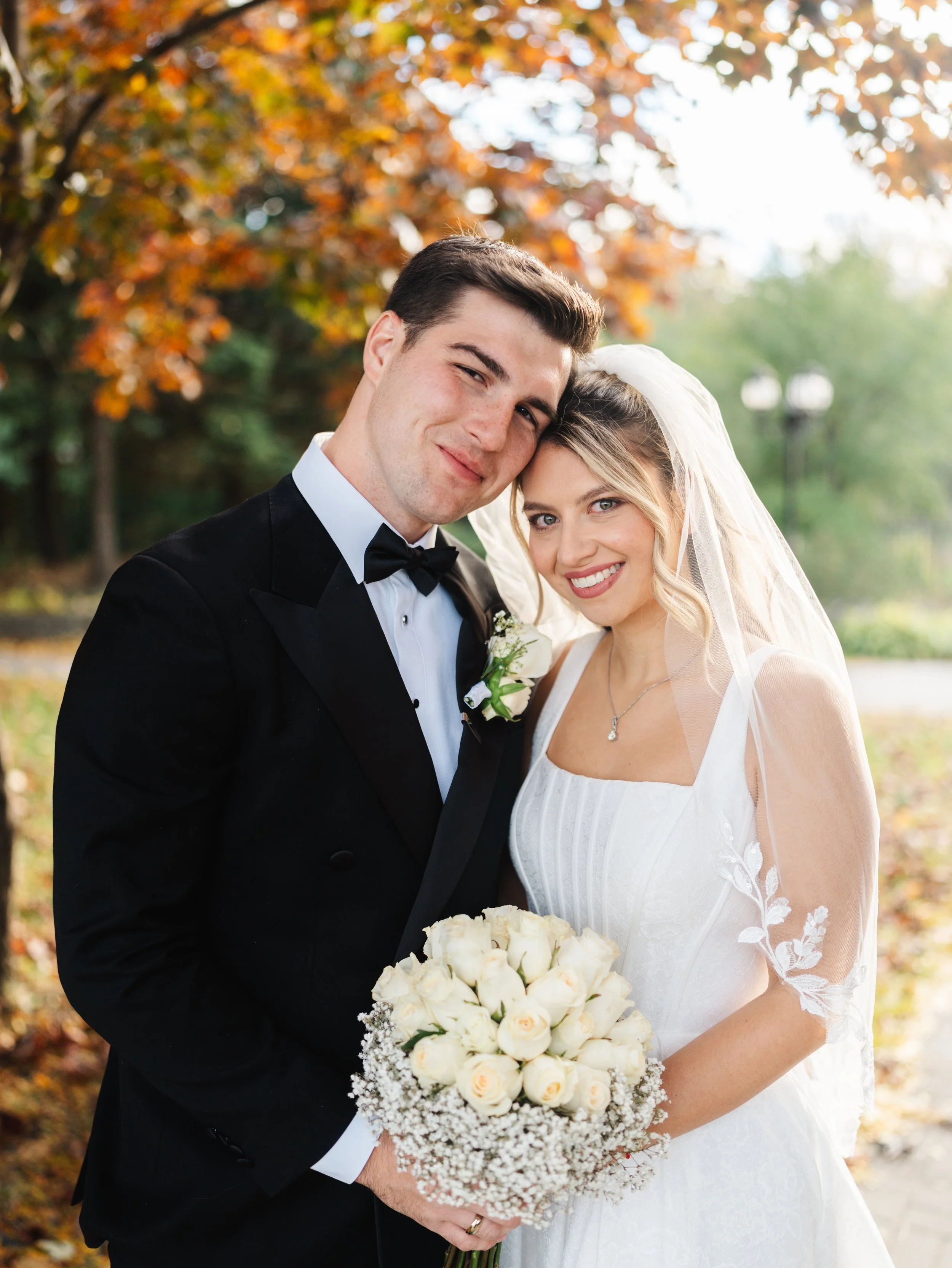 A bride and groom embrace outdoors during their wedding, with colorful autumn trees in the background. The bride is holding a bouquet of white roses and is smiling at the camera; the groom is smiling and leaning his face against hers.