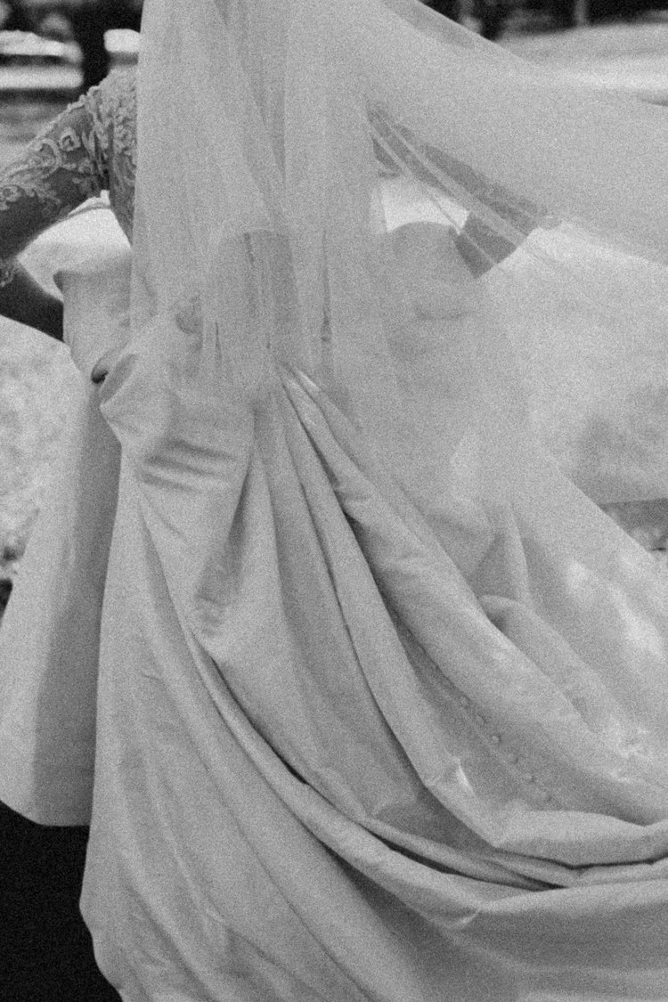 A person wearing a white dress with lace details on the sleeve, holding a bouquet of white flowers, and standing outside near a car.