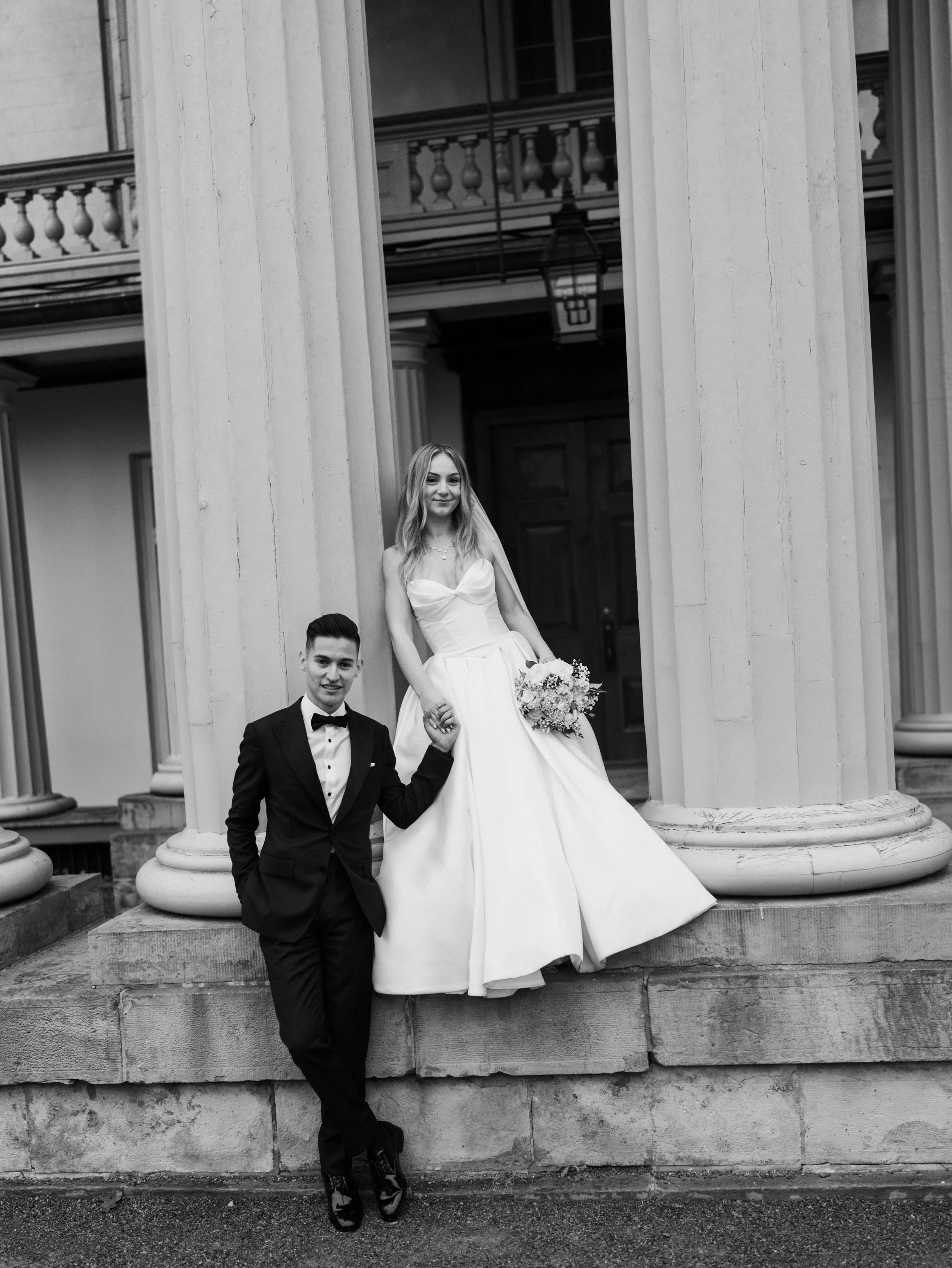 Black and white photograph of a bride and groom standing outside a classical building with columns. The bride is holding a bouquet and wearing a sleeveless wedding dress, while the groom is in a tuxedo. They are holding hands, smiling, and posing for