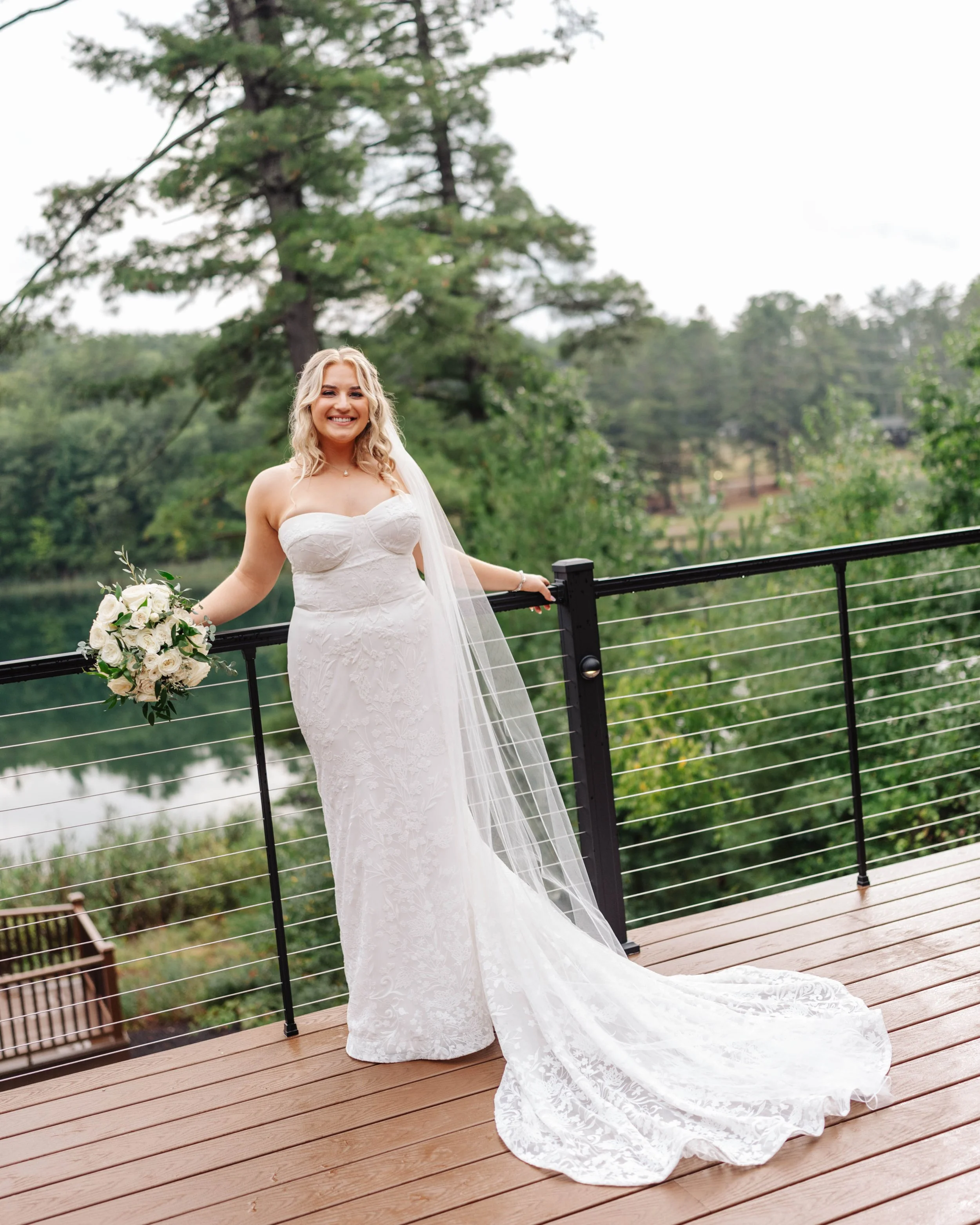 A bride in a white lace wedding dress with a long train, holding a bouquet of white roses, standing on a wooden deck near a lake with trees in the background, smiling.
