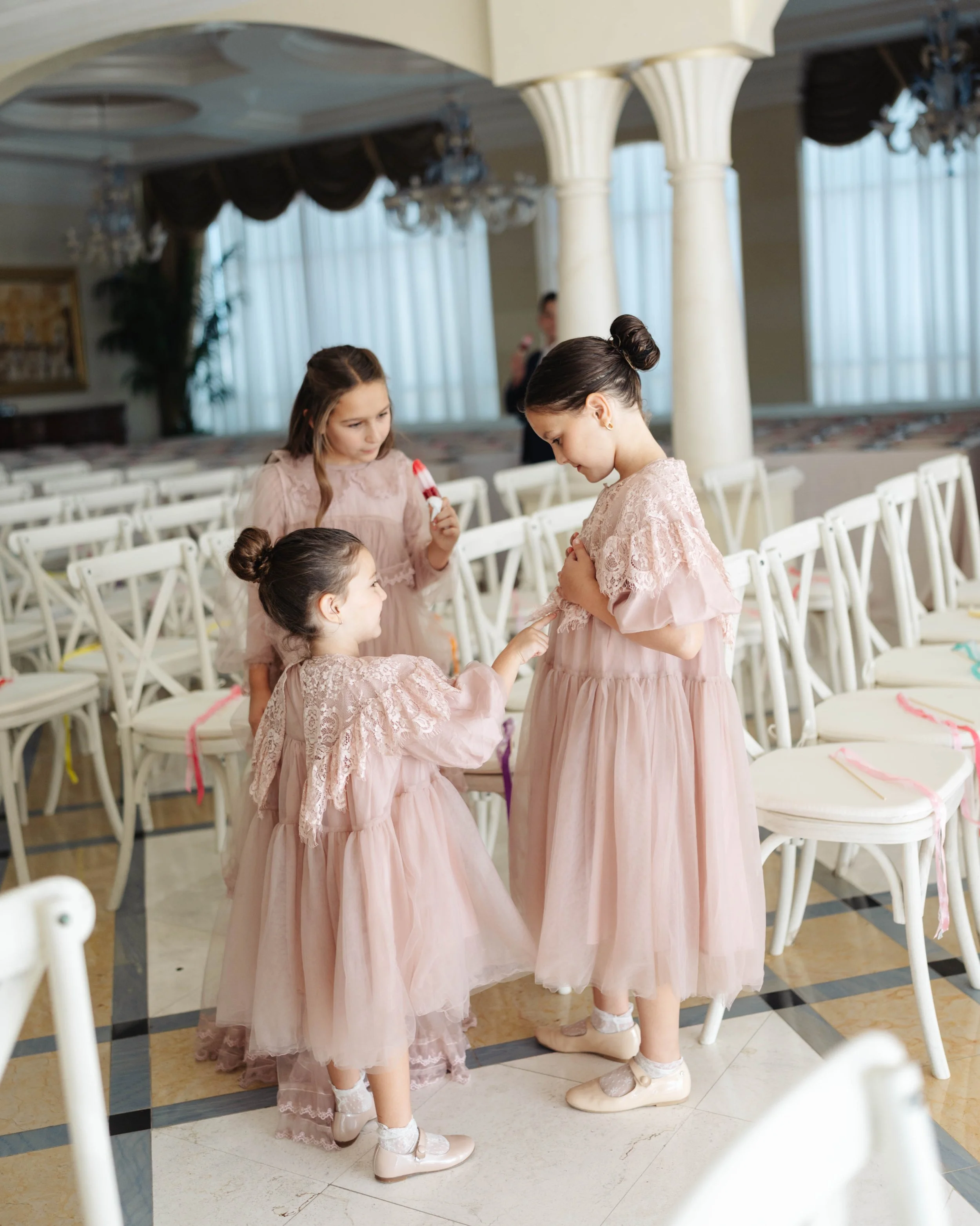 Three young girls dressed in pink lace dresses and ballet shoes, two children are standing, one is kneeling, in an indoor event space with many empty chairs decorated with pink, purple, and yellow ribbons.