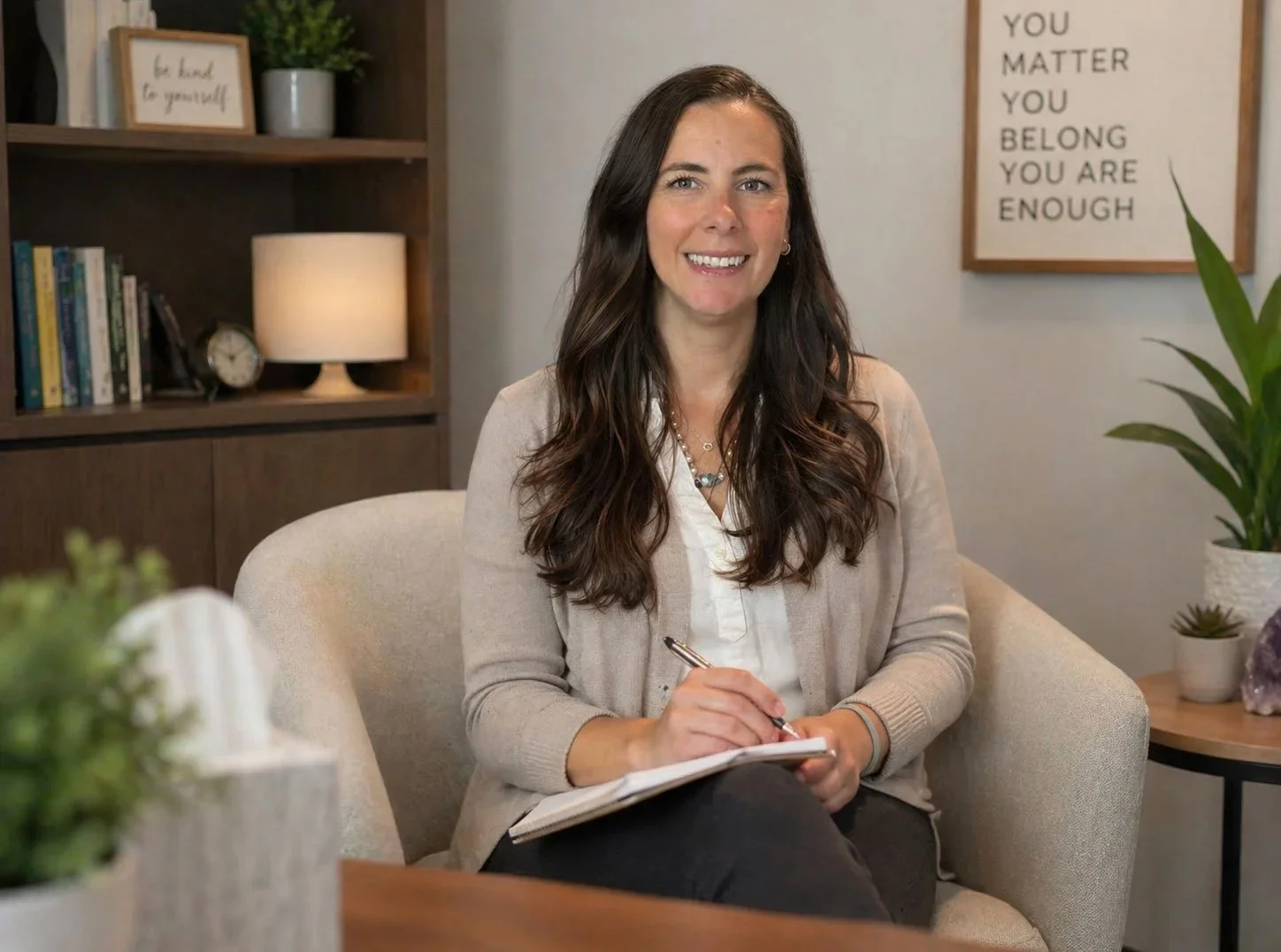 Woman sitting on a beige armchair, smiling, and writing in a notebook in a cozy room with a bookshelf, a lamp, a framed motivational quote on the wall, and houseplants.