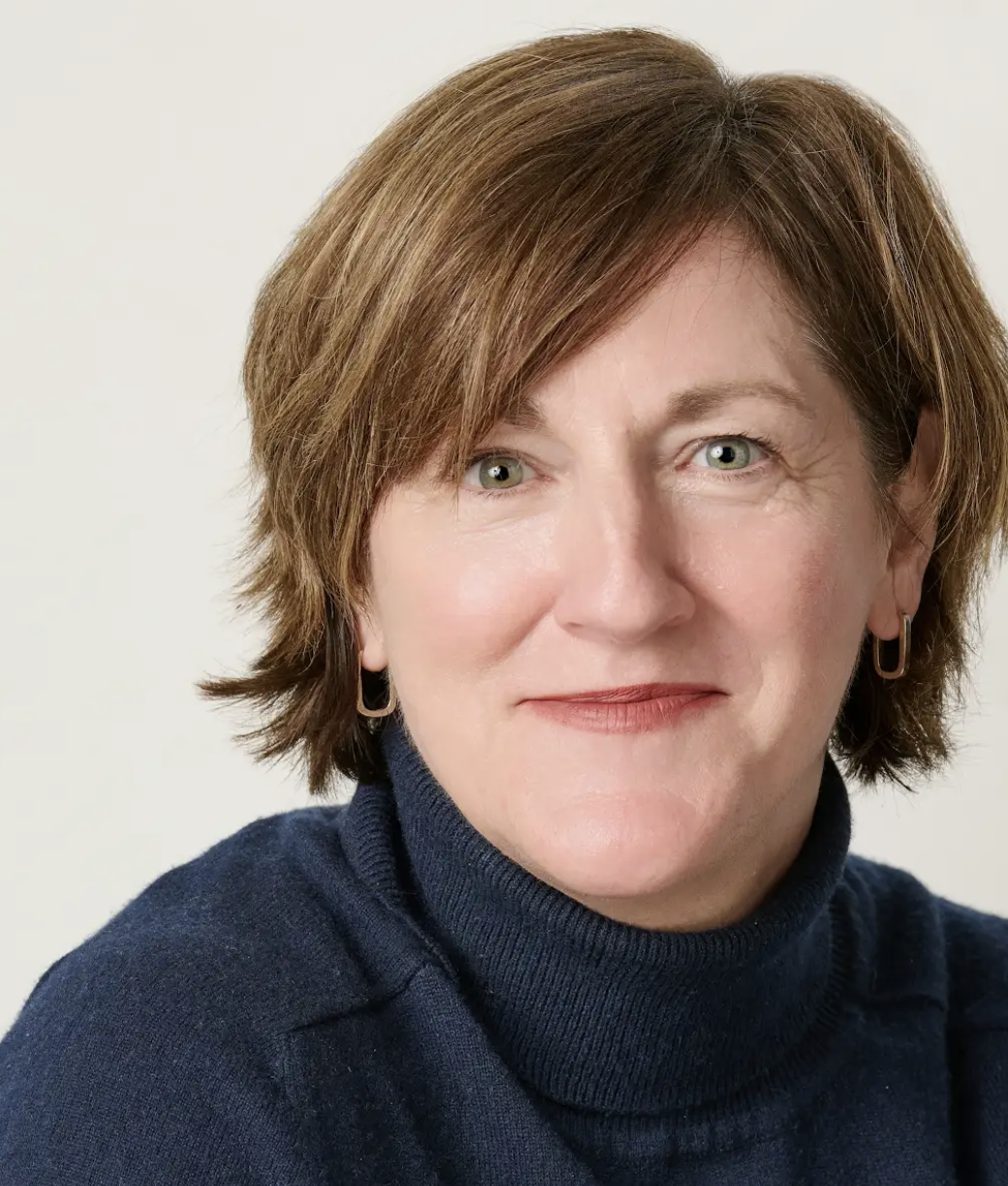 A woman with short, reddish-brown hair, wearing silver earrings and a navyblue turtleneck sweater, looking directly at the camera with a neutral expression.