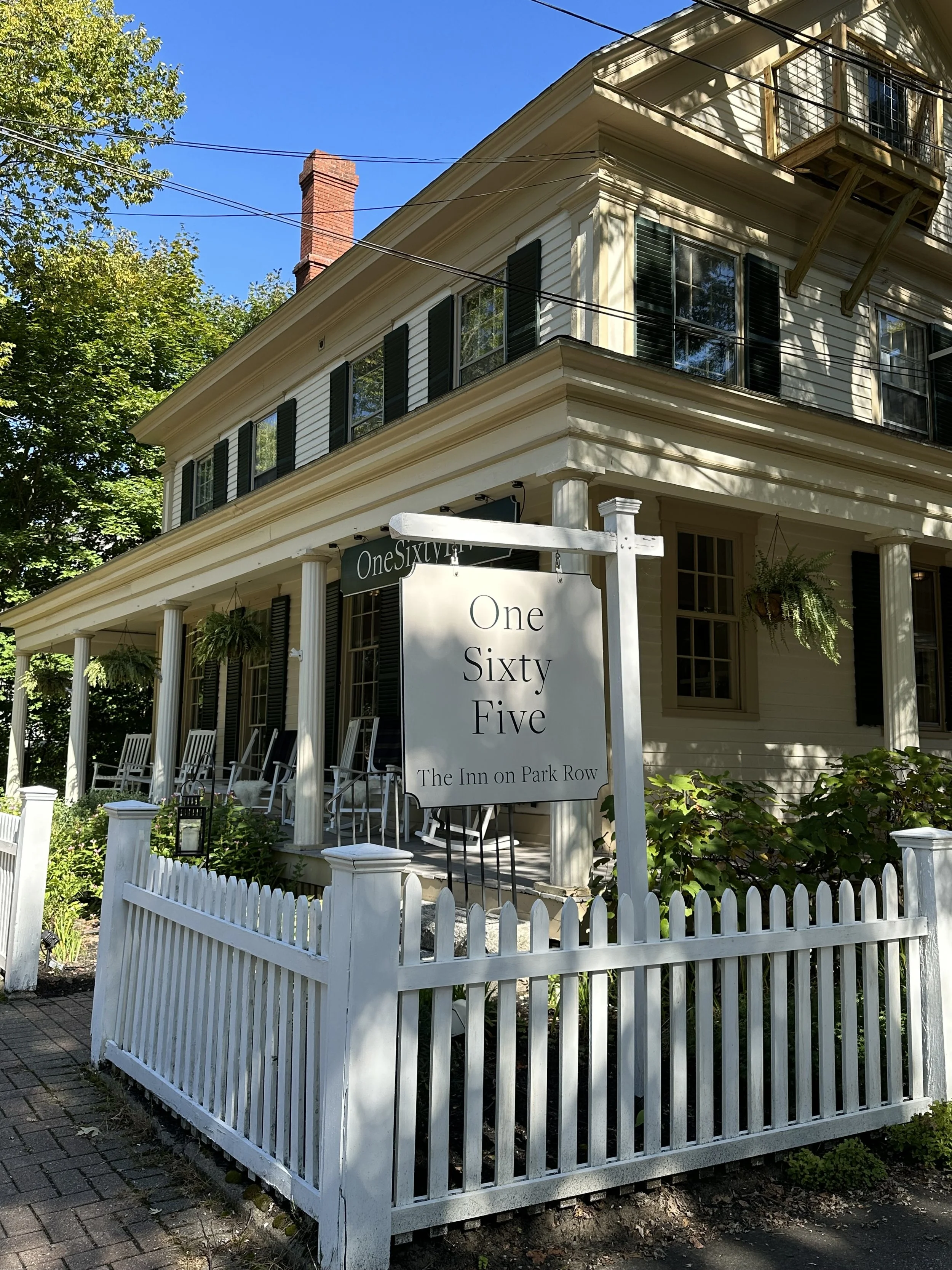 A historic, cream-colored house with black shutters, a porch with white rocking chairs, and hanging plants. There is a white picket fence and a sign that says 'One Sixty Five, The Inn on Park Row'.