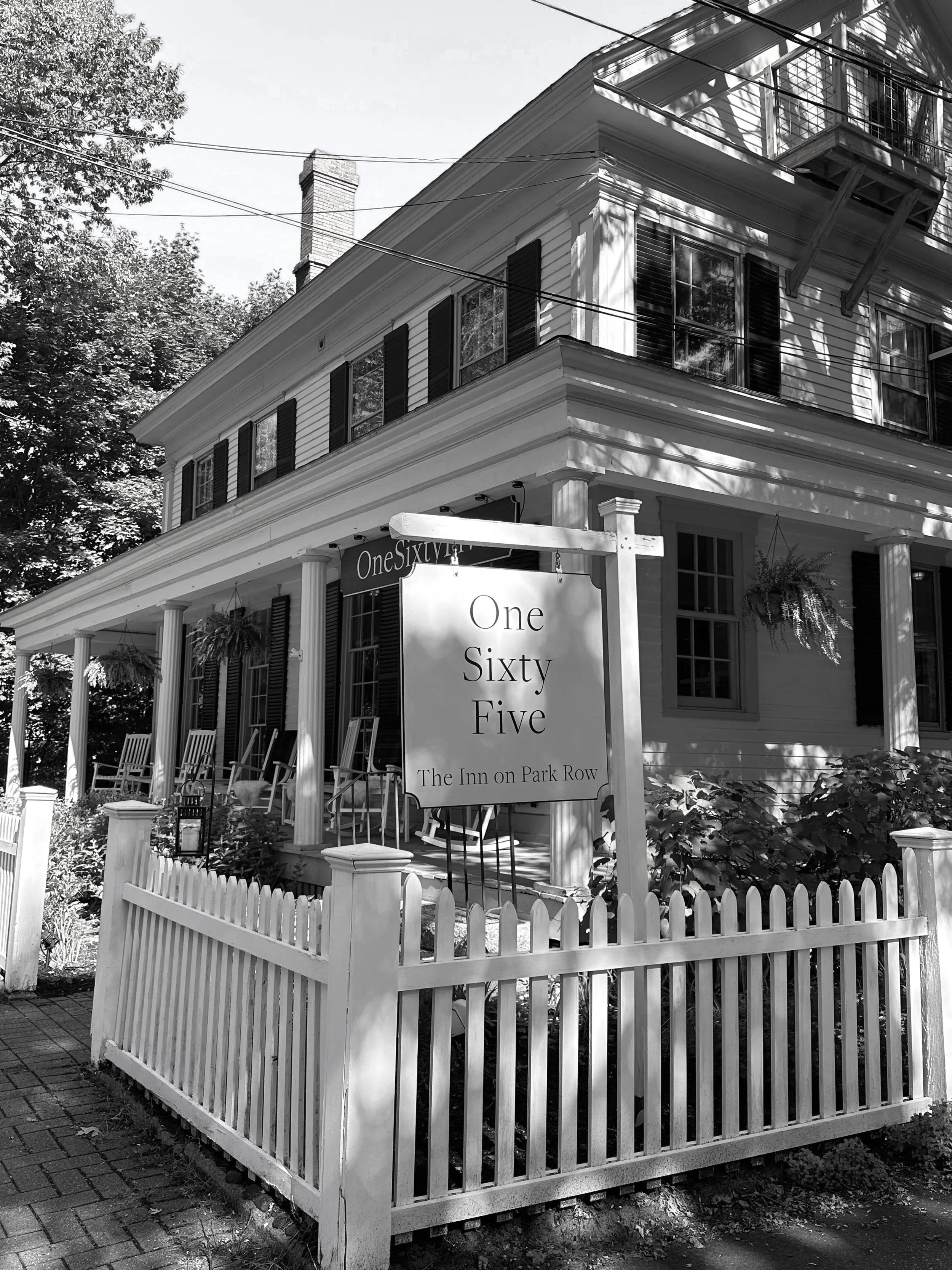 A black and white photo of a large, two-story house with a porch, black shutters, and hanging plants. There is a white picket fence in front with a sign that reads 'One Sixty Five' and 'The Inn on Park Row'.