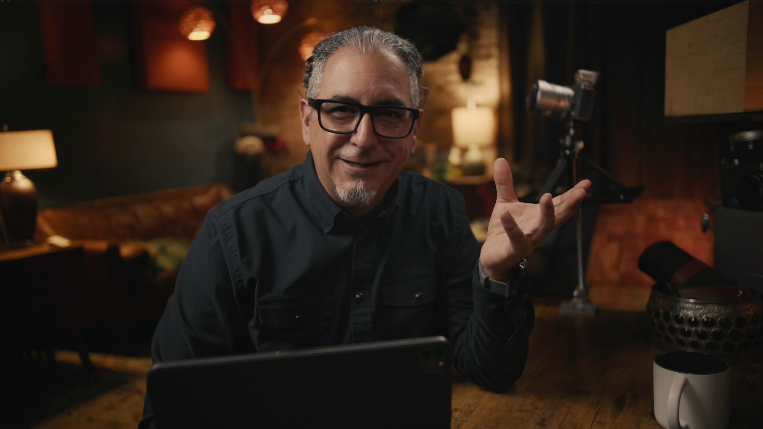 A man with gray hair, glasses, and a goatee sitting at a wooden table in a dimly lit room, smiling and gesturing with one hand, with cameras and equipment nearby.