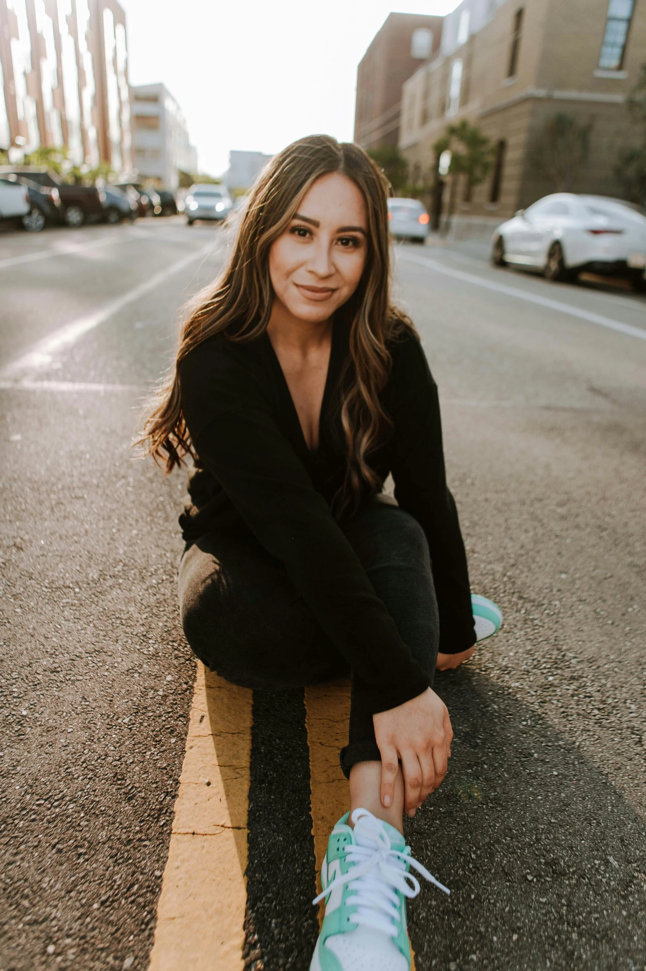 A woman in black clothing squats on a street, holding her ankle, with a smile, in an urban area with parked cars and buildings in the background.