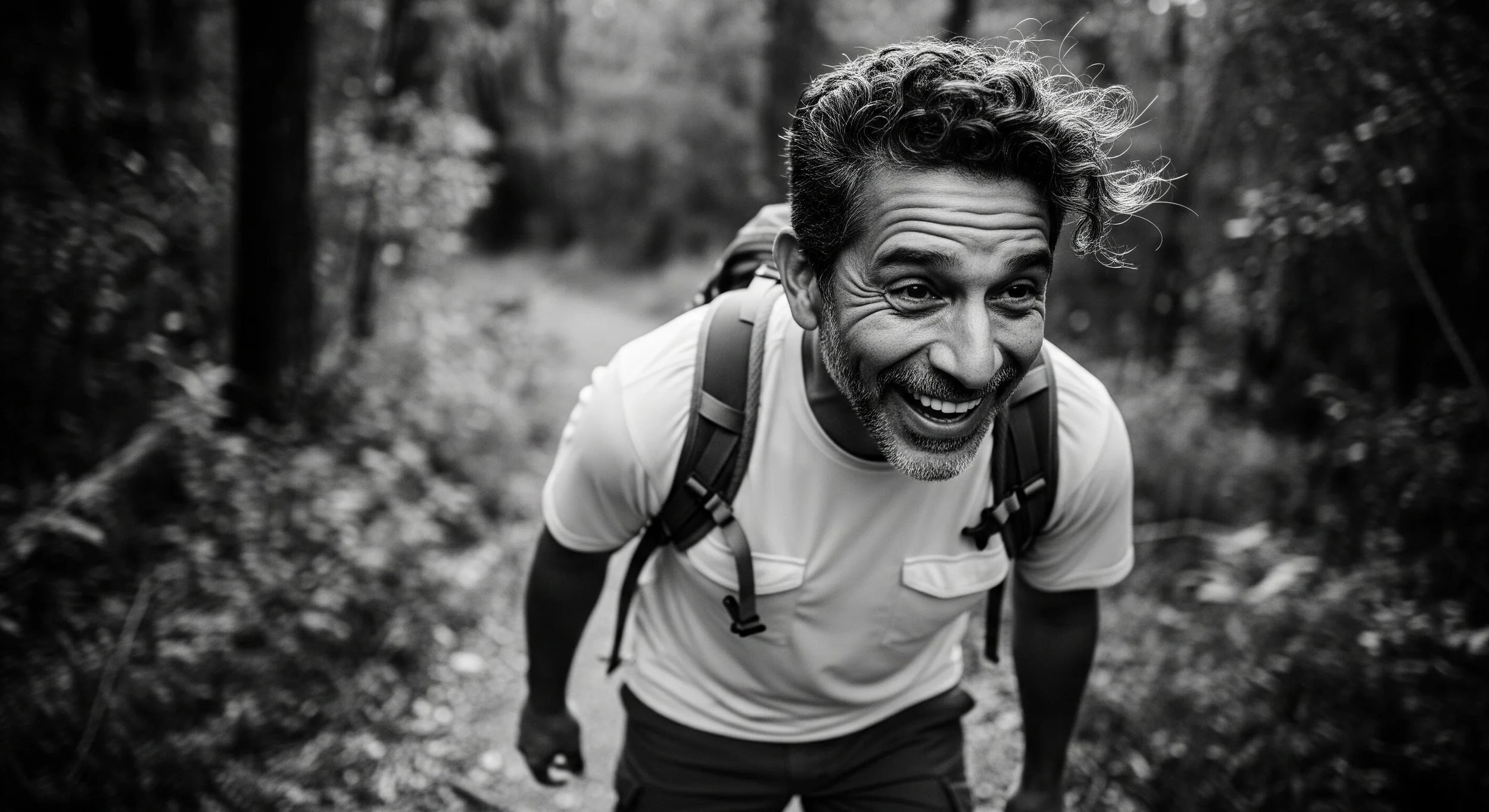 A man with curly hair and a beard is hiking on a forest trail, smiling and leaning forward with his hands on his knees, wearing a backpack.