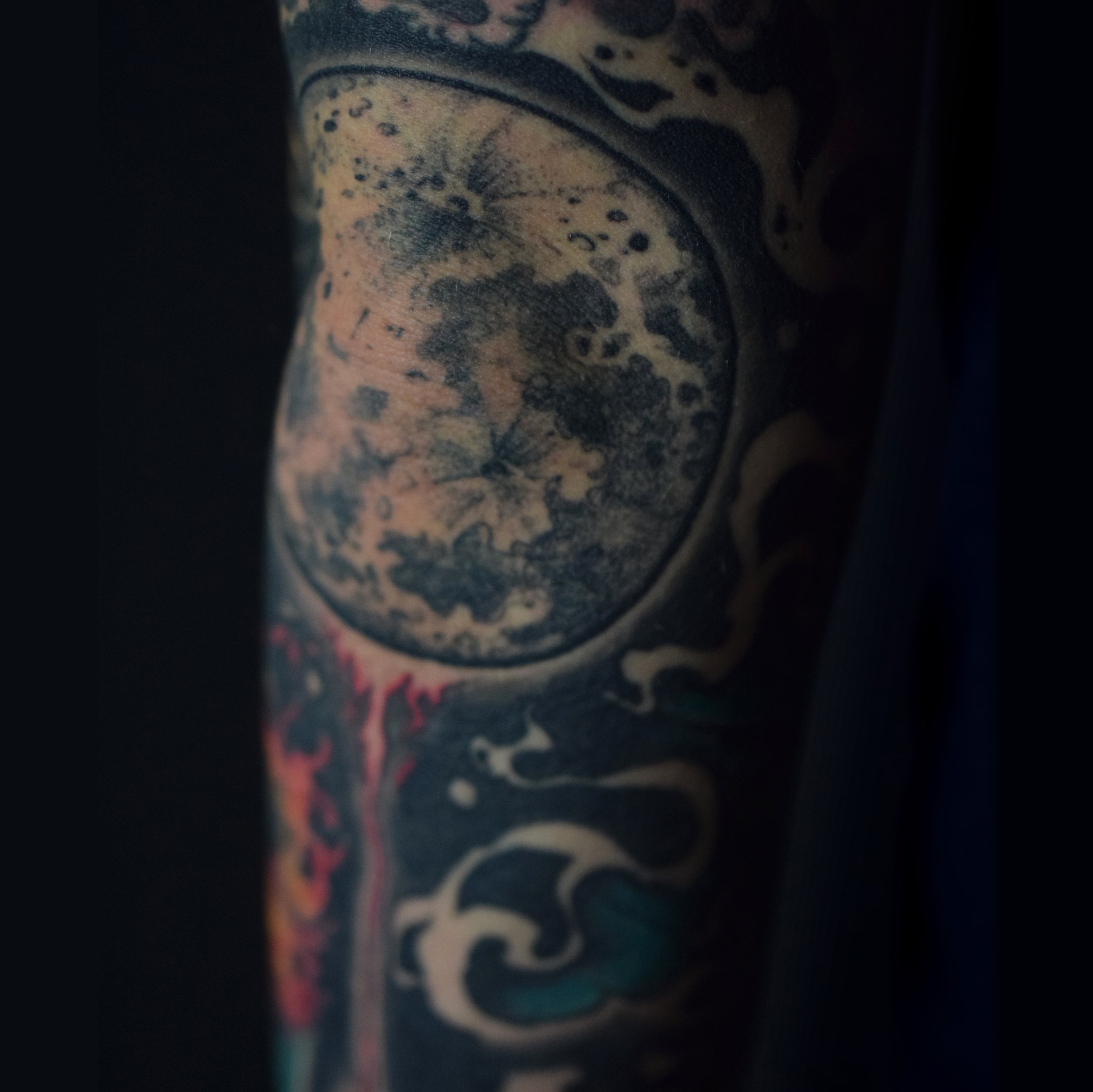 Close-up of a tattoo featuring a detailed moon with clouds and celestial elements surrounded by decorative patterns on a person's arm.