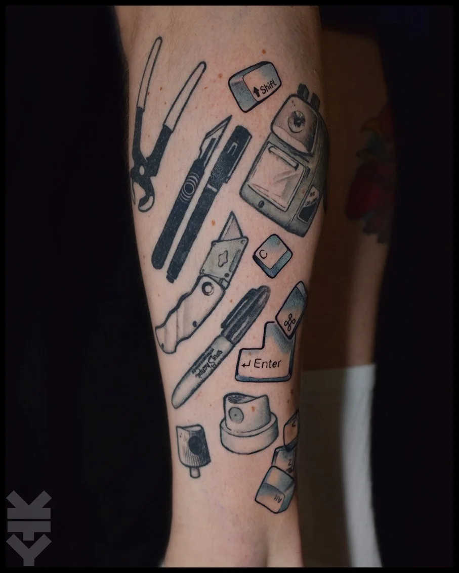 Tattoo of various technology and computer-related objects on a person's arm, including a pair of pliers, a pen, a USB flash drive, a computer monitor, keyboard keys with 'Shift' and 'Enter', a mouse, a speaker, and other electronic accessories.